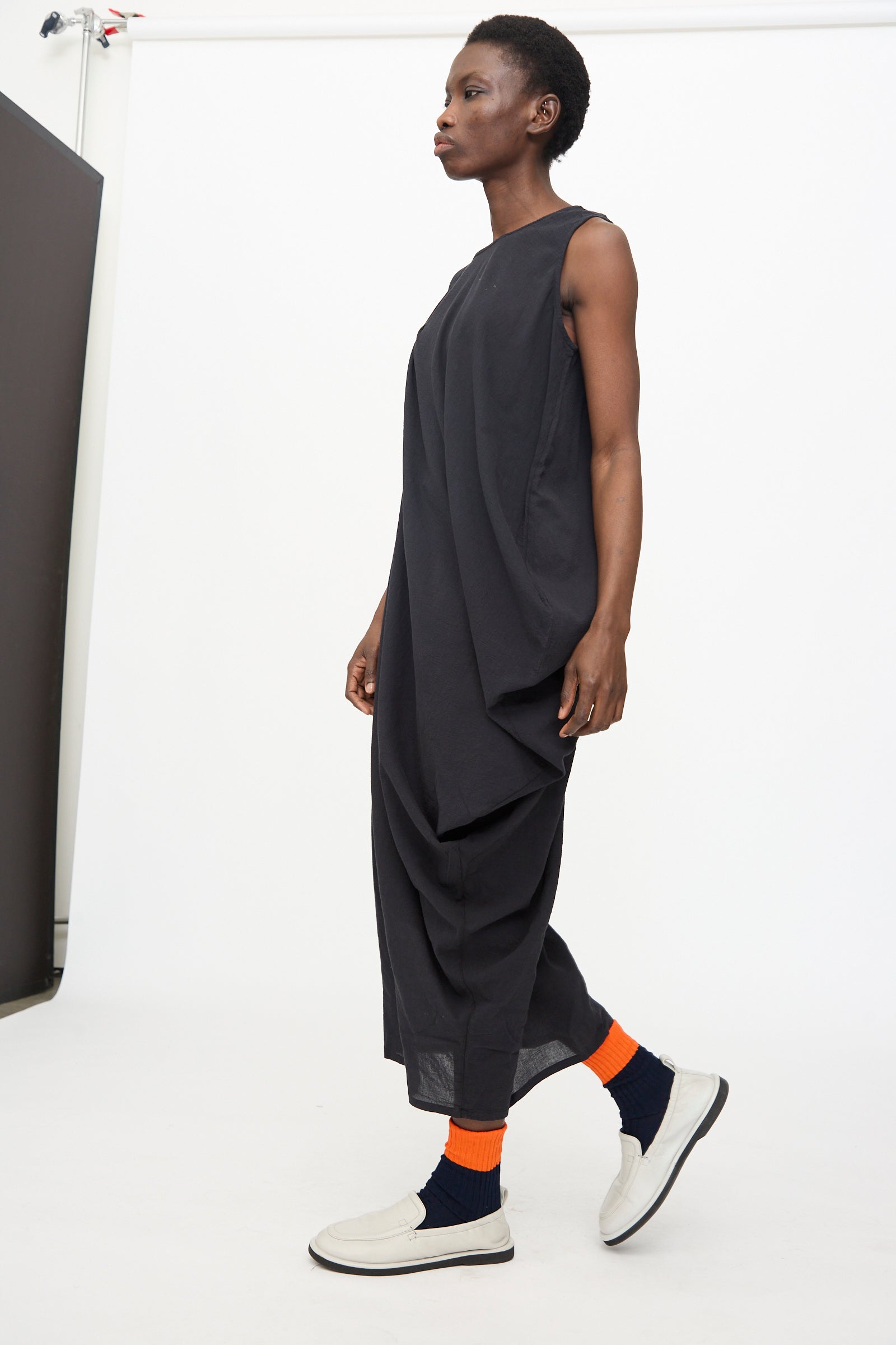A person in the Black Crane Greek Column Dress in Sumi Black, paired with orange and navy socks and white slip-on shoes, stands against a plain white background.