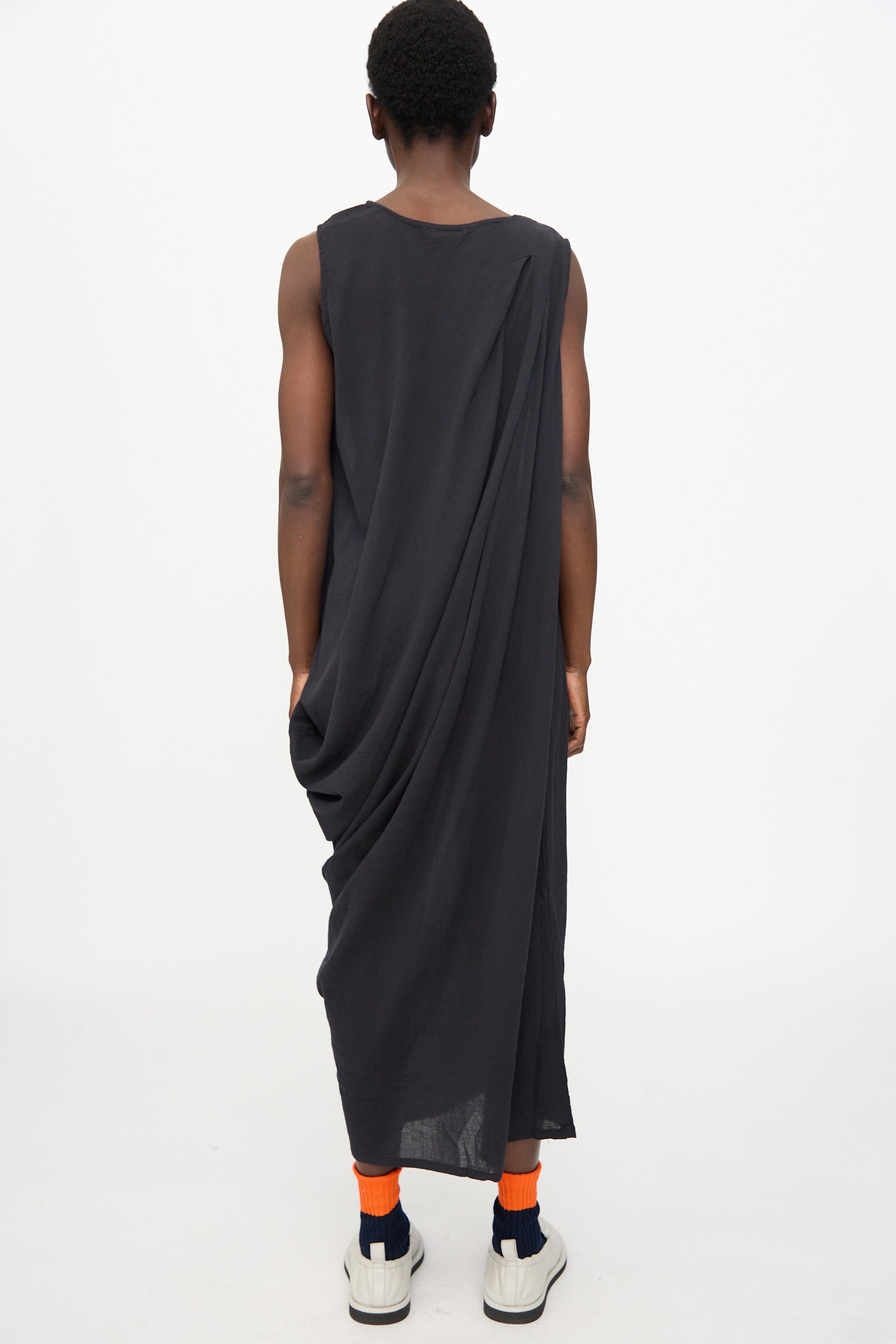 A person faces away from the camera against a plain background, wearing Black Crane's Greek Column Dress in Sumi Black, paired with orange socks and white sneakers. The dress has a relaxed fit and sleeveless cotton gauze design.