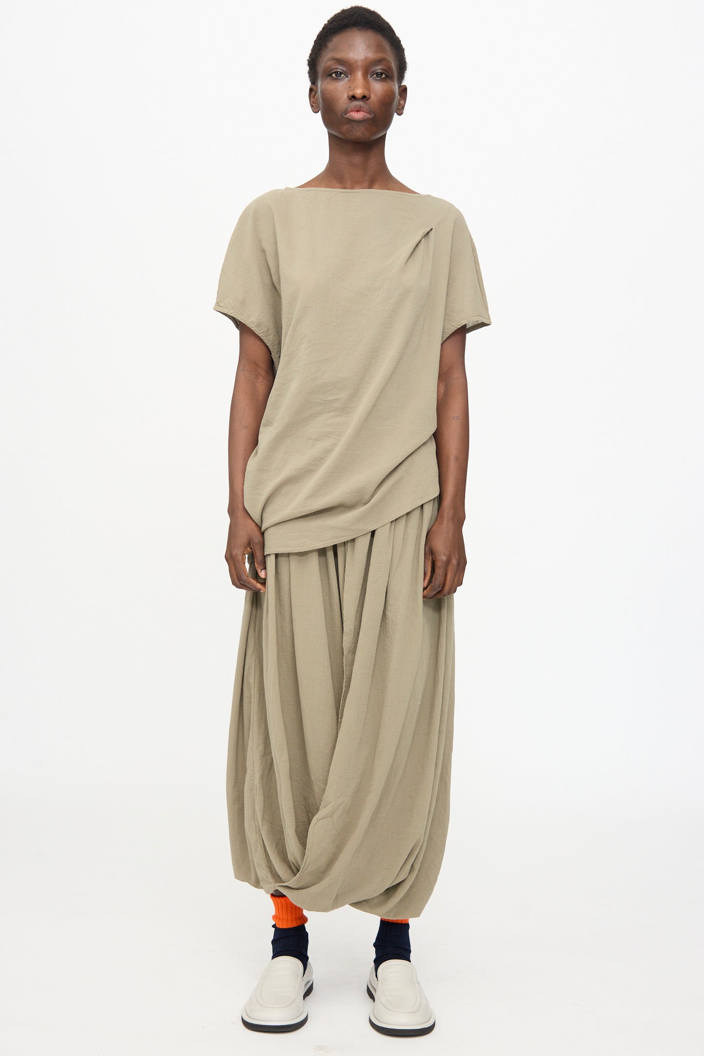 A person wears the Black Crane Greek Column Skirt in Cement with a loose beige top, orange socks, and white slip-on shoes, standing against a plain white background.