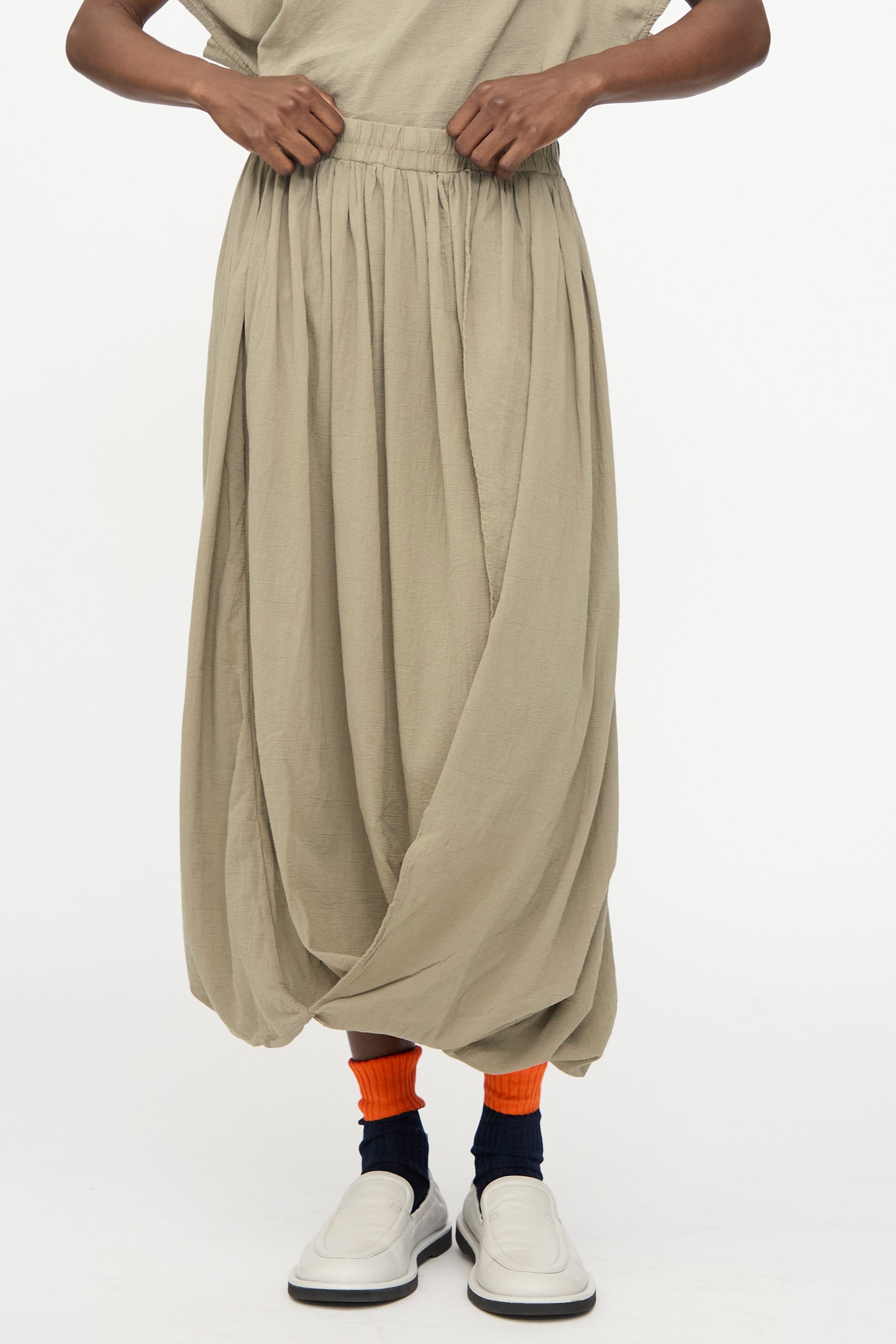 A person wears the Black Crane Greek Column Skirt in Cement, featuring a loose fit and elastic waistband, paired with orange and black socks and white slip-on shoes against a plain background.