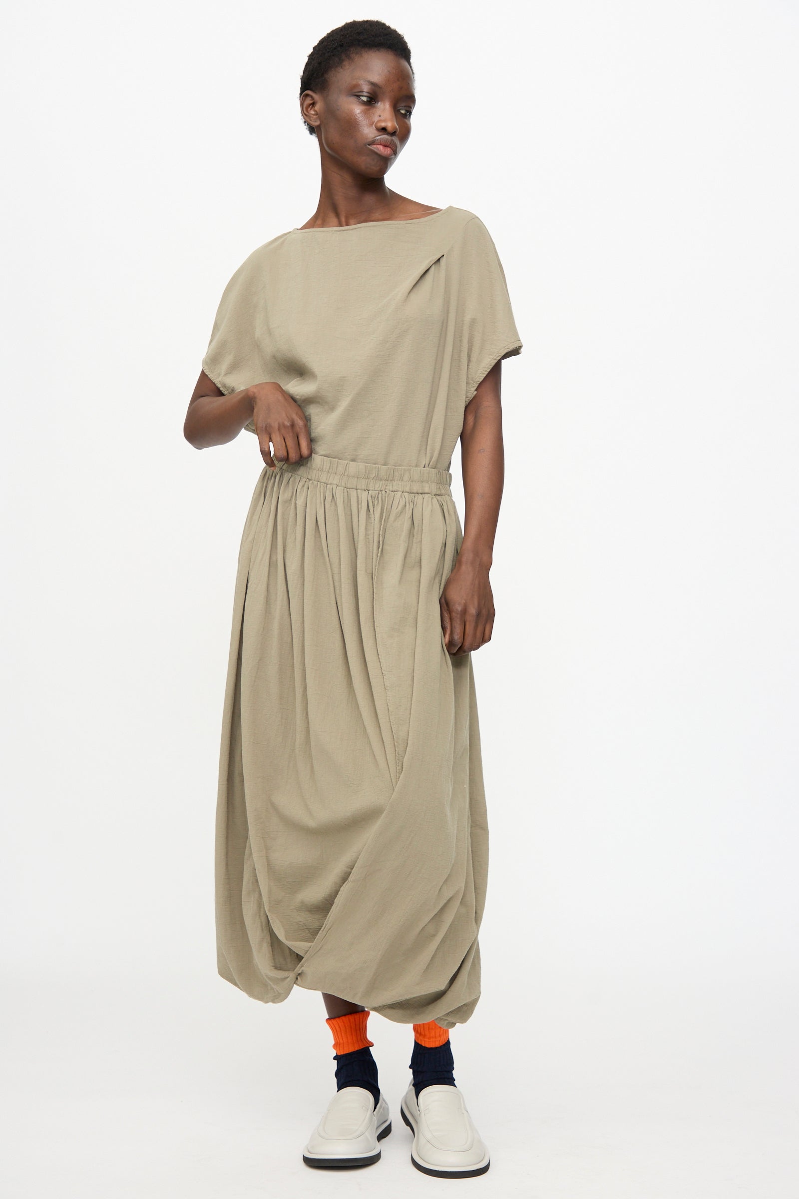 Person wearing the Black Crane Greek Column Skirt in Cement, paired with orange socks, navy ankle covers, and white slip-on shoes, standing against a plain white background.