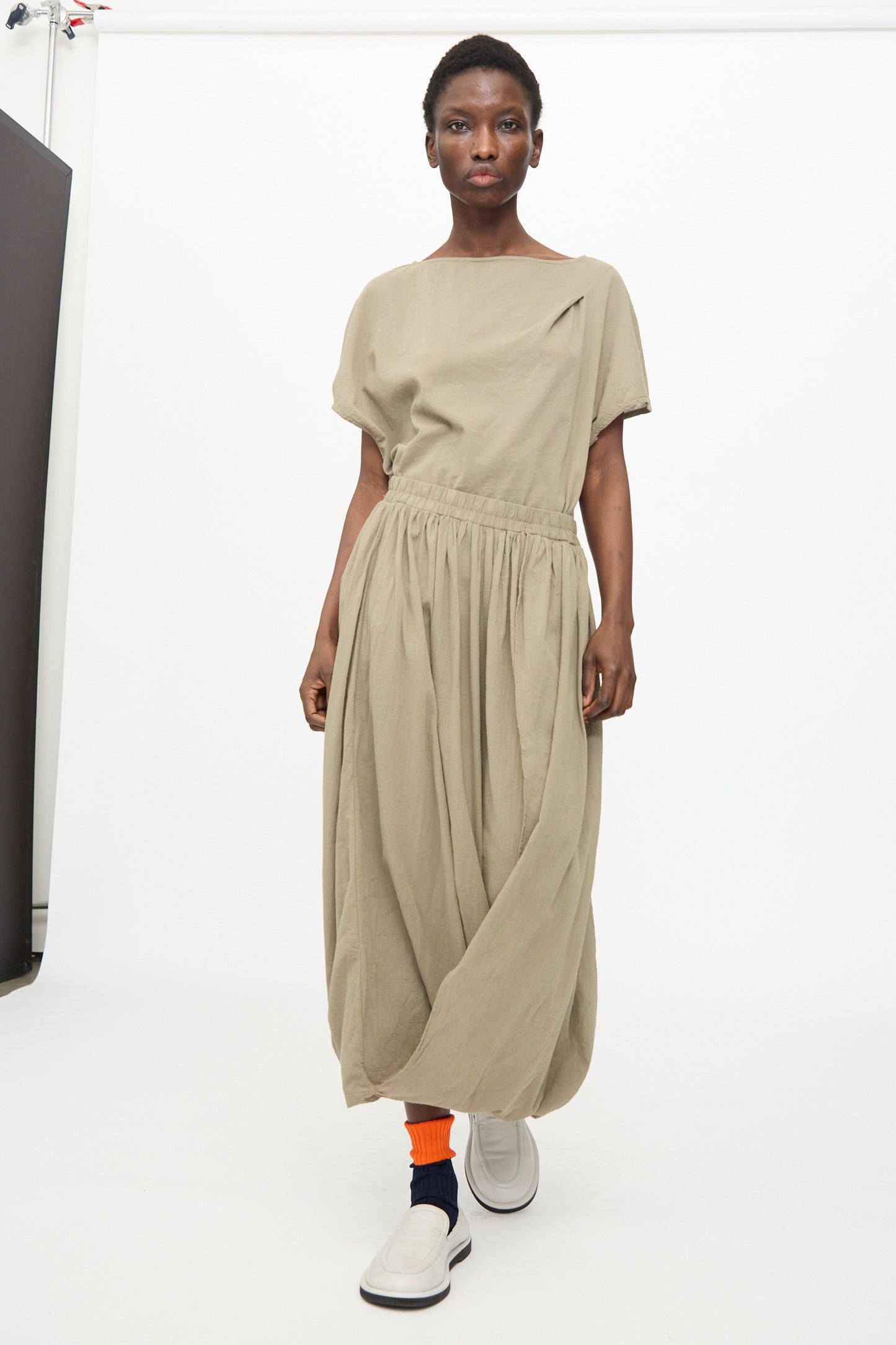 Person wearing the Black Crane Greek Column Skirt in Cement, paired with orange socks and white slip-on shoes, standing against a plain studio background.