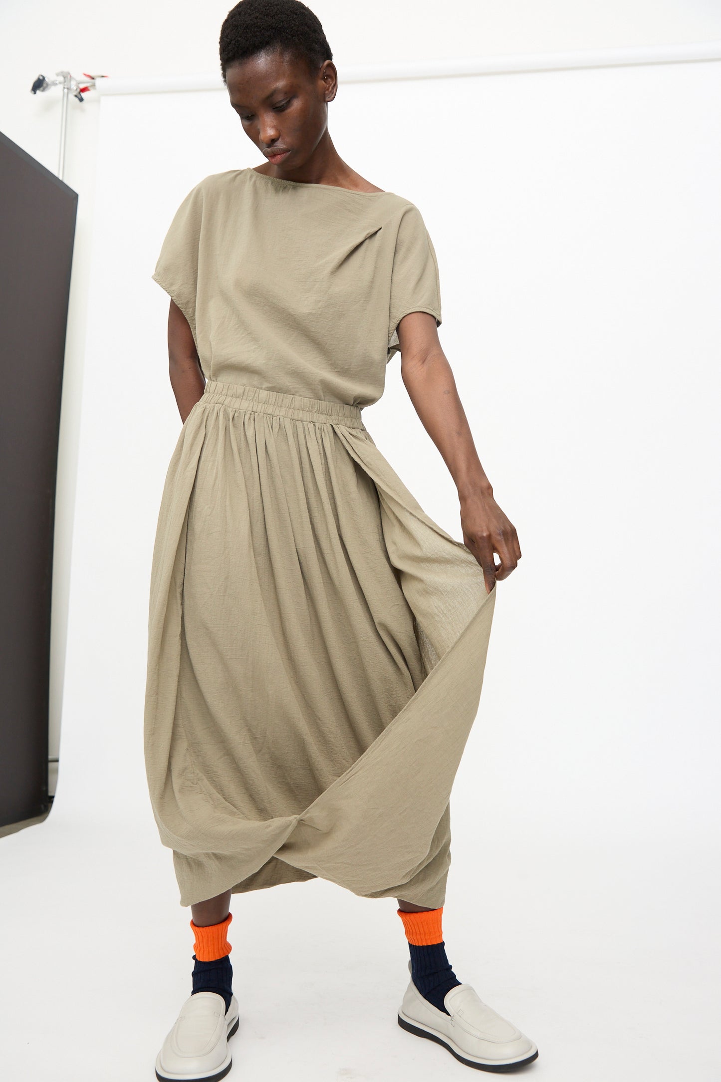 Model wears the Black Crane Greek Column Skirt in Cement with a matching top, orange and navy socks, and white slip-on shoes, highlighting a relaxed fit against a plain white background.