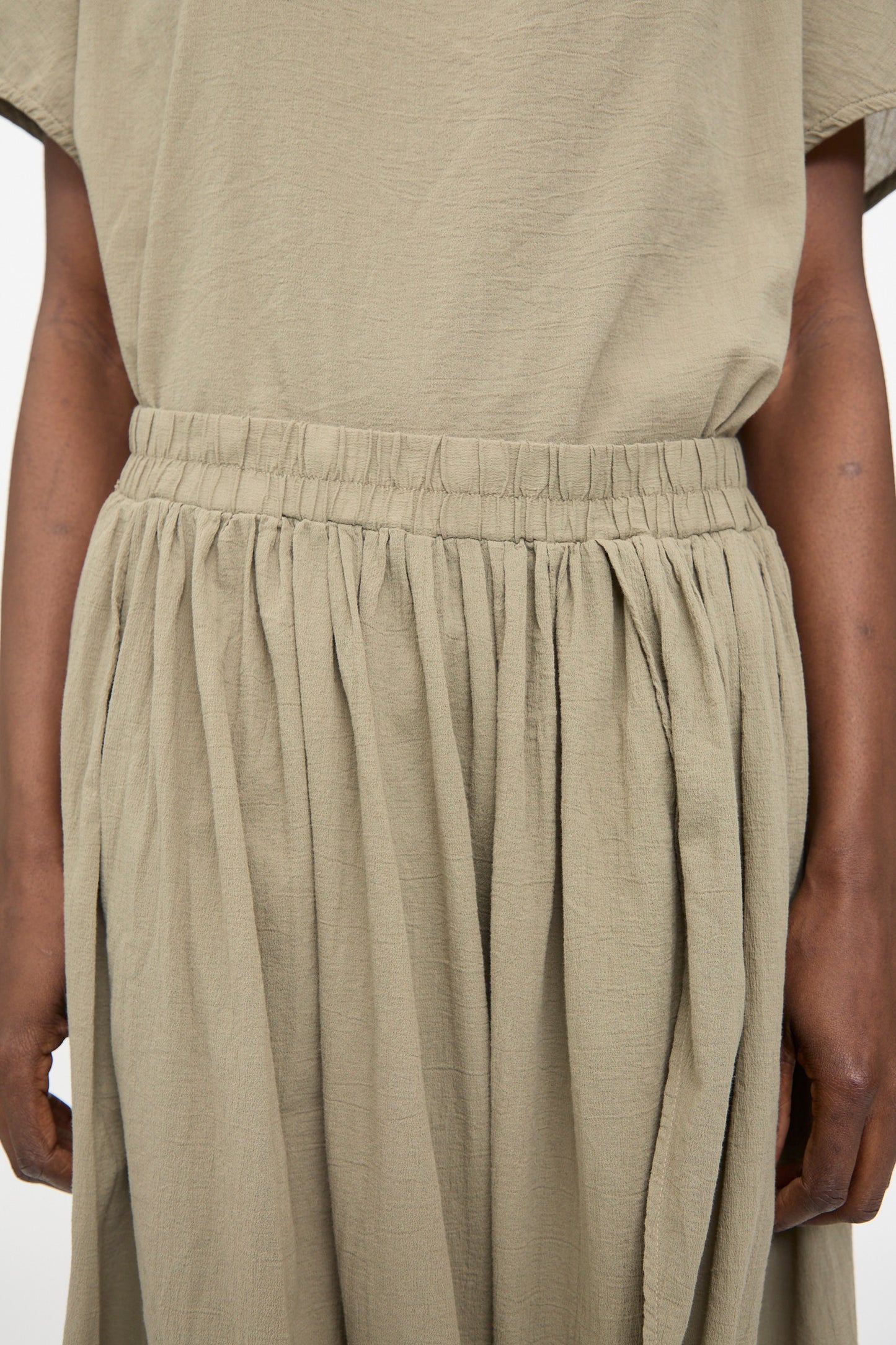 A person wearing the Black Crane Greek Column Skirt in Cement, featuring a relaxed fit with a gathered elastic waistband. Only the torso and part of the arms are visible.