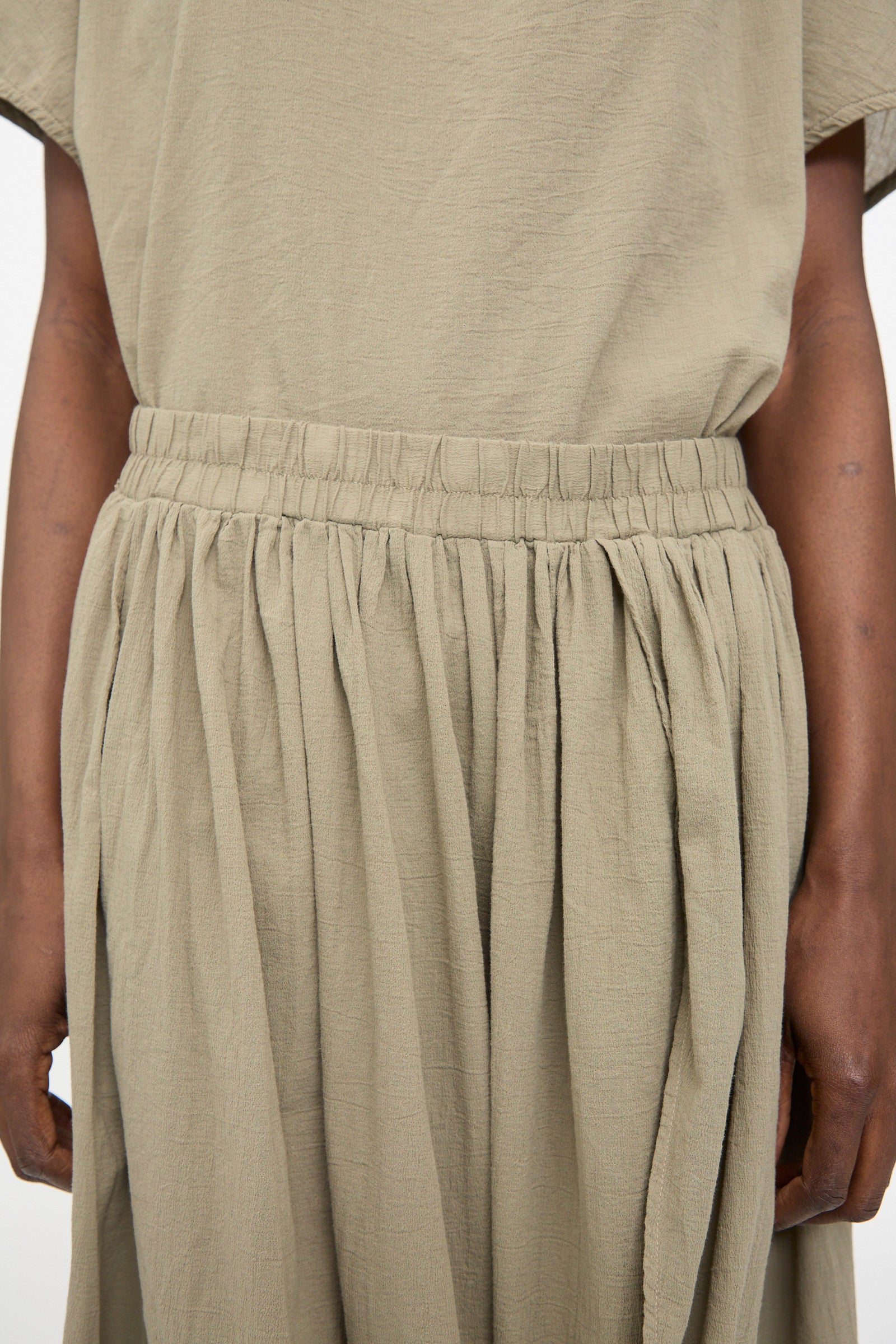 A person wearing the Black Crane Greek Column Skirt in Cement, featuring a relaxed fit with a gathered elastic waistband. Only the torso and part of the arms are visible.