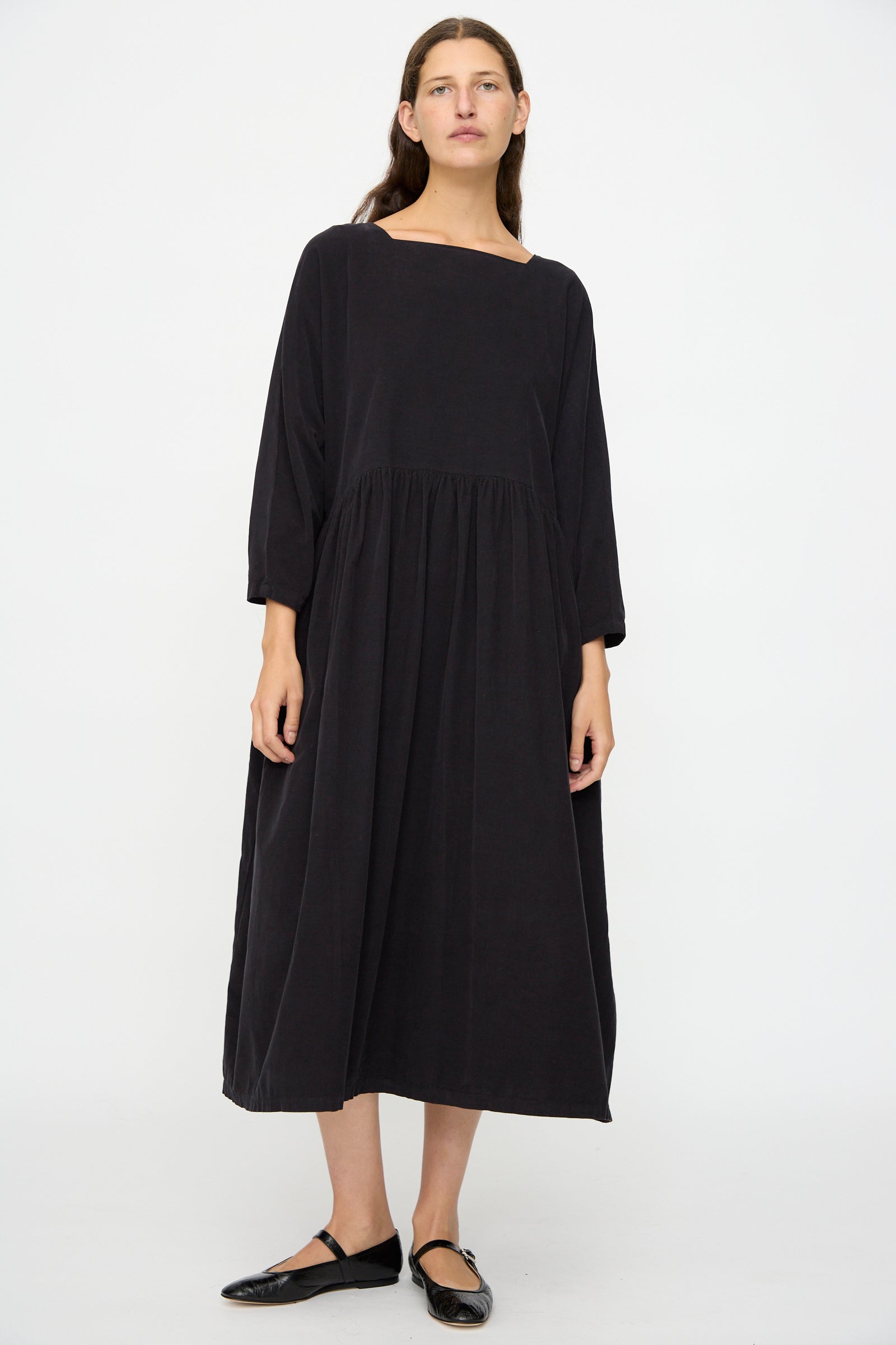 A person stands against a white background wearing the oversized fit, long-sleeved Tradi Dress in Dark Navy by Black Crane and black flat shoes.