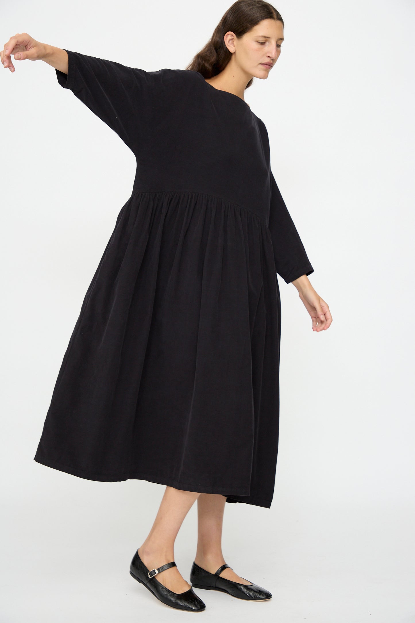 A woman stands with one arm raised, wearing the Black Crane Tradi Dress in Dark Navy—an oversized fit, long-sleeved organic cotton midi dress—and black flat shoes against a plain white background.
