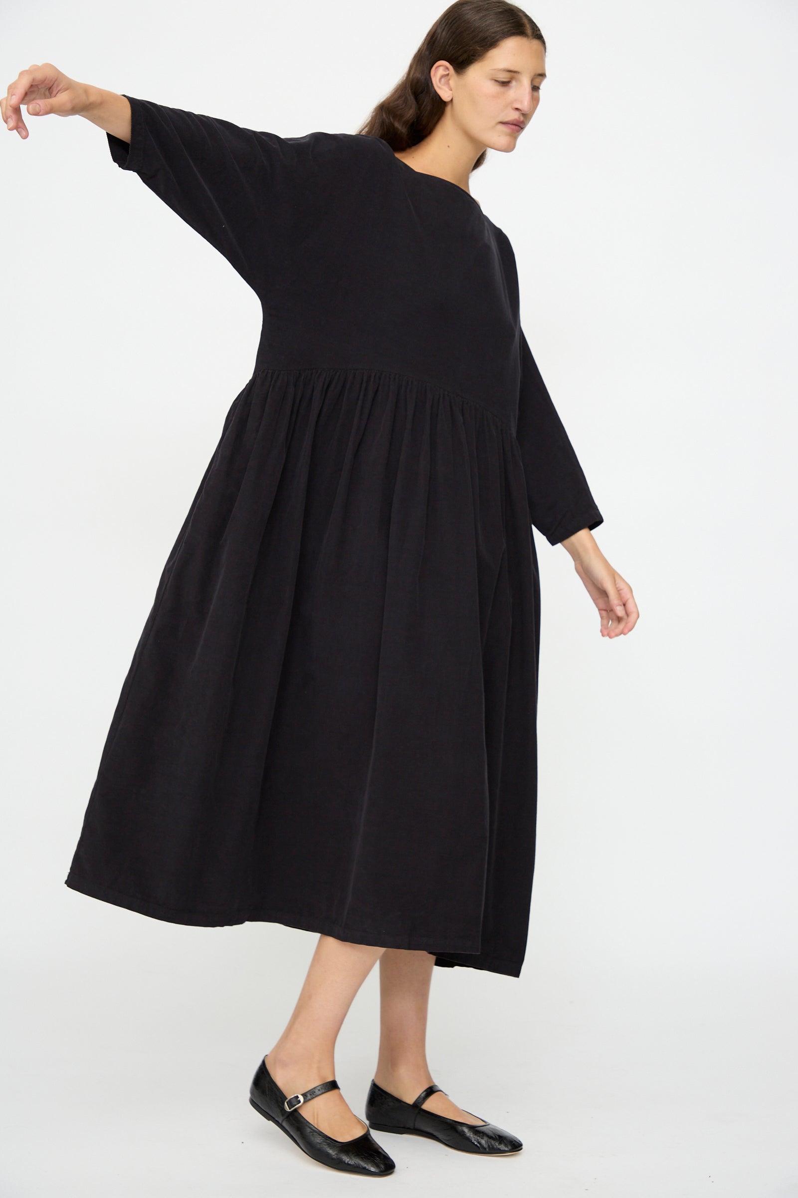 A woman stands with one arm raised, wearing the Black Crane Tradi Dress in Dark Navy—an oversized fit, long-sleeved organic cotton midi dress—and black flat shoes against a plain white background.