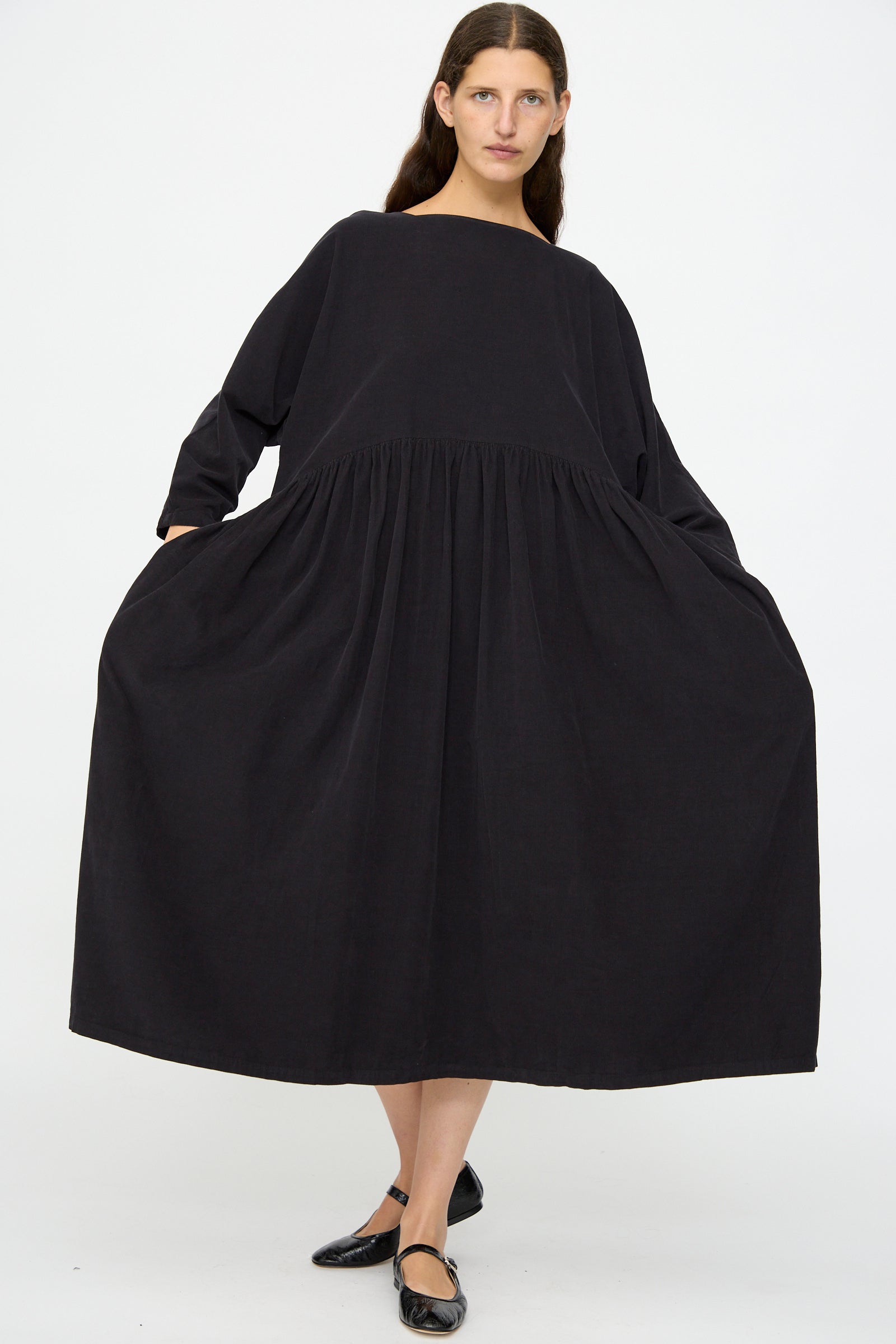 A person stands against a white background wearing the Black Crane Tradi Dress in Dark Navy—an oversized, long-sleeve, organic cotton midi dress—and black flats, holding out each side of the dress.