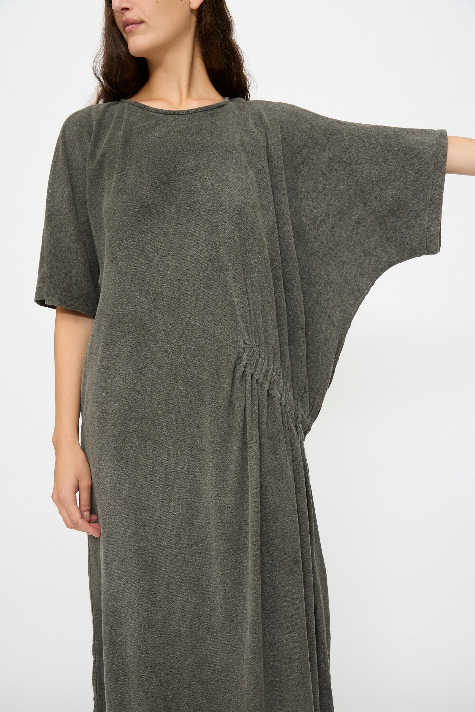 A person wears the Black Crane Greek Dress in Slate—a relaxed-fit, short-sleeved grey dress with waist gathers, made from a soft cotton-rayon blend—standing against a plain white background.