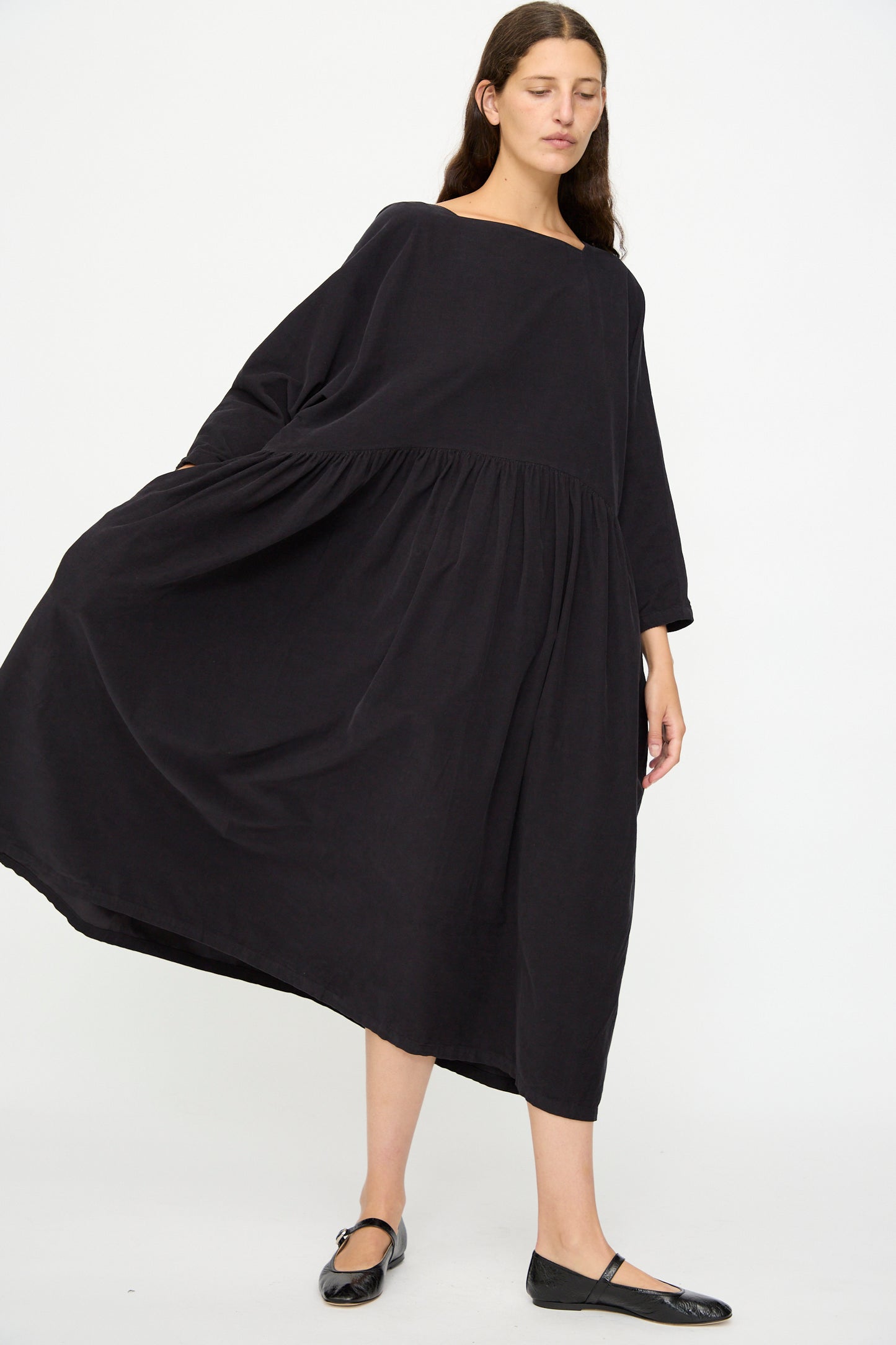 A woman stands against a plain white background wearing the Black Crane Tradi Dress in Dark Navy, an oversized long dress with long sleeves, paired with black flats.