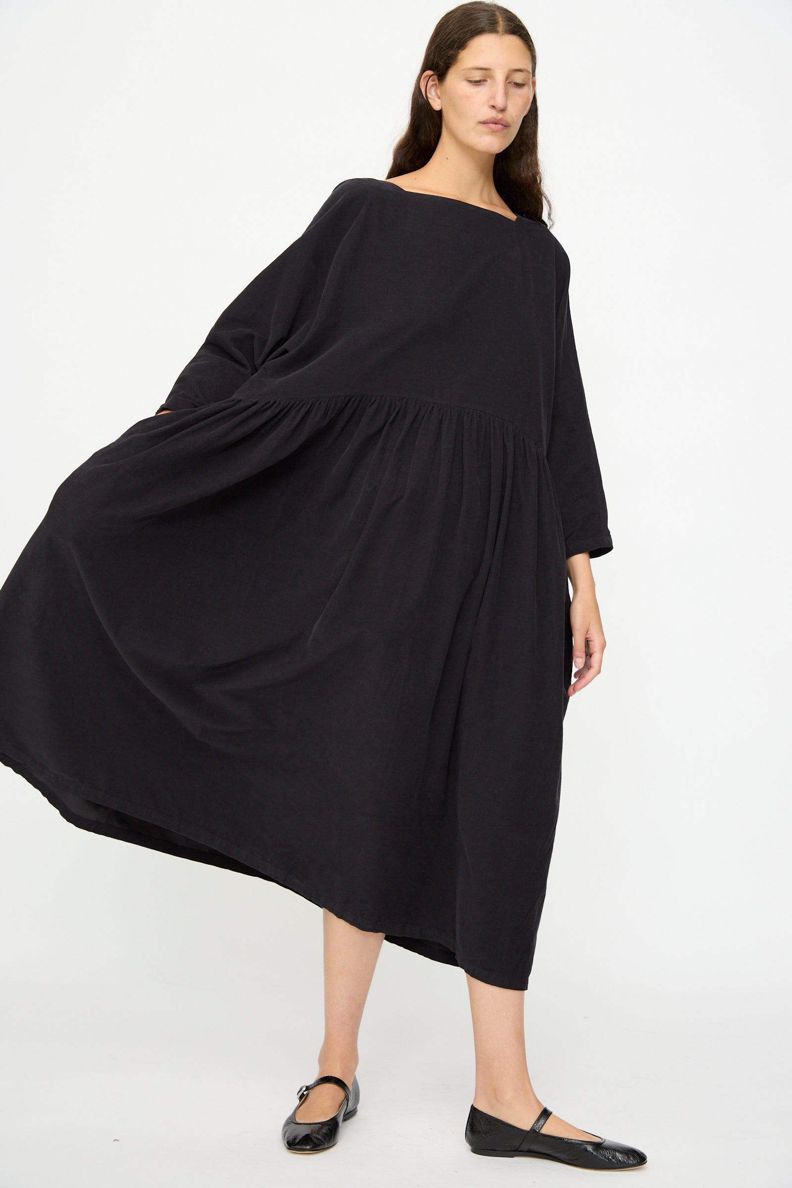 A woman stands against a plain white background wearing the Black Crane Tradi Dress in Dark Navy, an oversized long dress with long sleeves, paired with black flats.