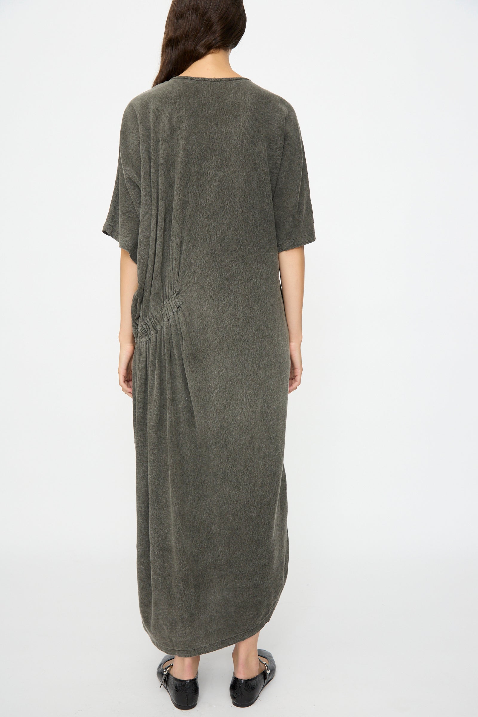 Person stands with back to camera wearing the Black Crane Greek Dress in Slate, a relaxed fit, ankle-length maxi with short sleeves and ruched side detail, paired with flat black shoes; hair over one shoulder.