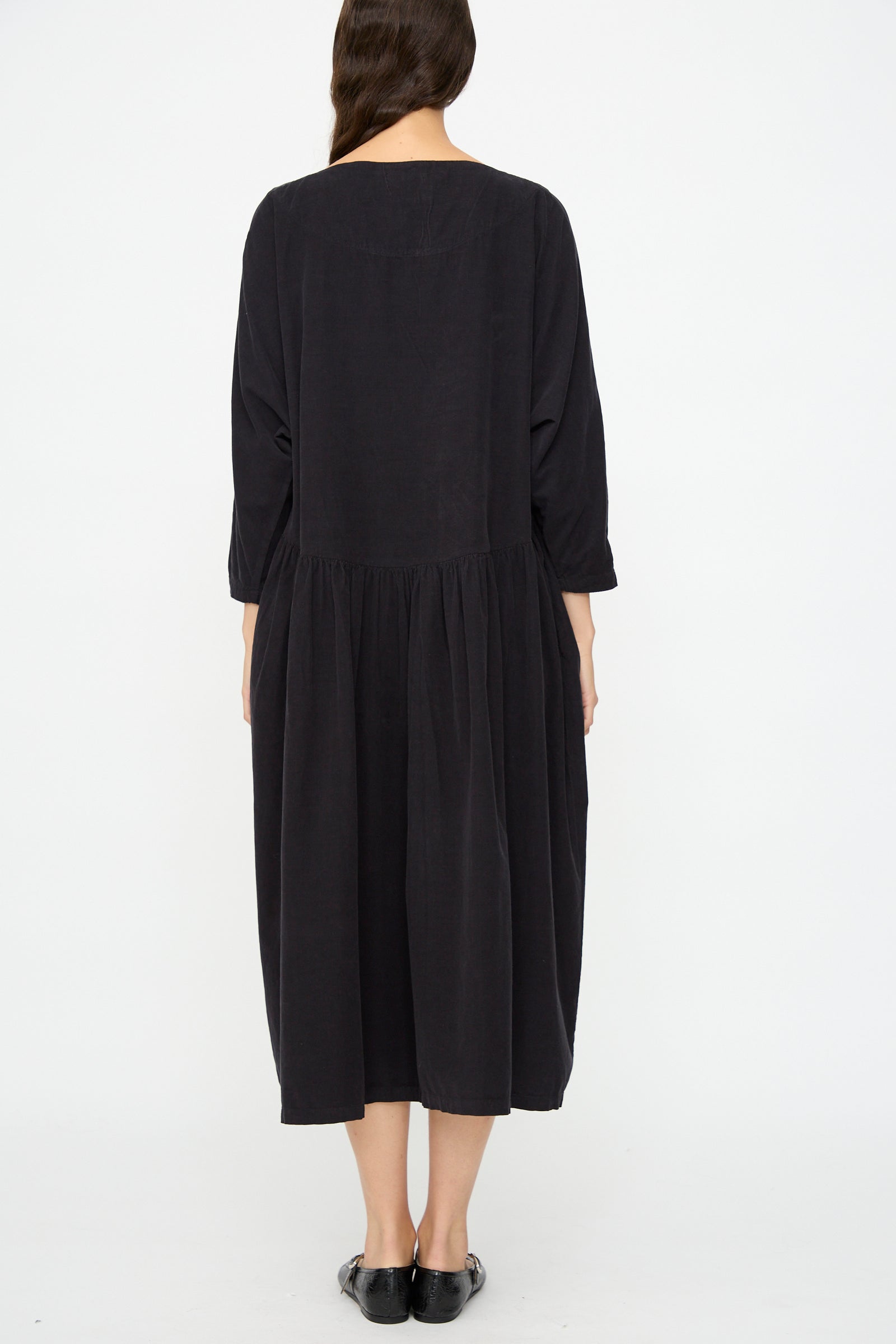 A person with long hair is seen from the back, wearing the Black Crane Tradi Dress in Dark Navy—an oversized, long-sleeved midi dress—with black shoes, against a plain white background.