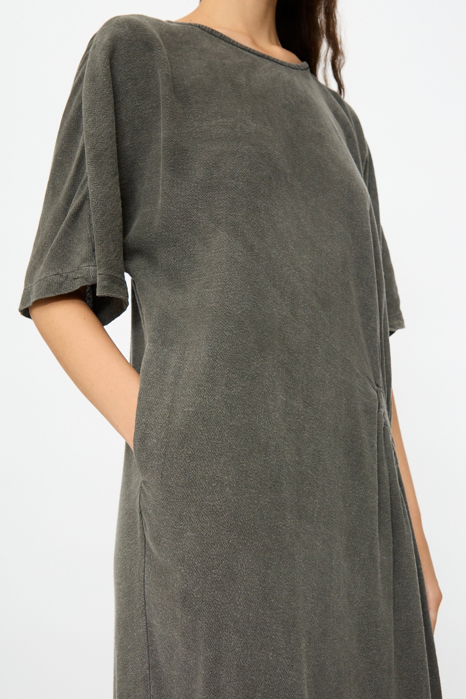 A person wears the Black Crane Greek Dress in Slate—a relaxed-fit, short-sleeve, dark gray cotton-rayon blend dress with side pockets—shown from shoulders to knees against a plain light background.