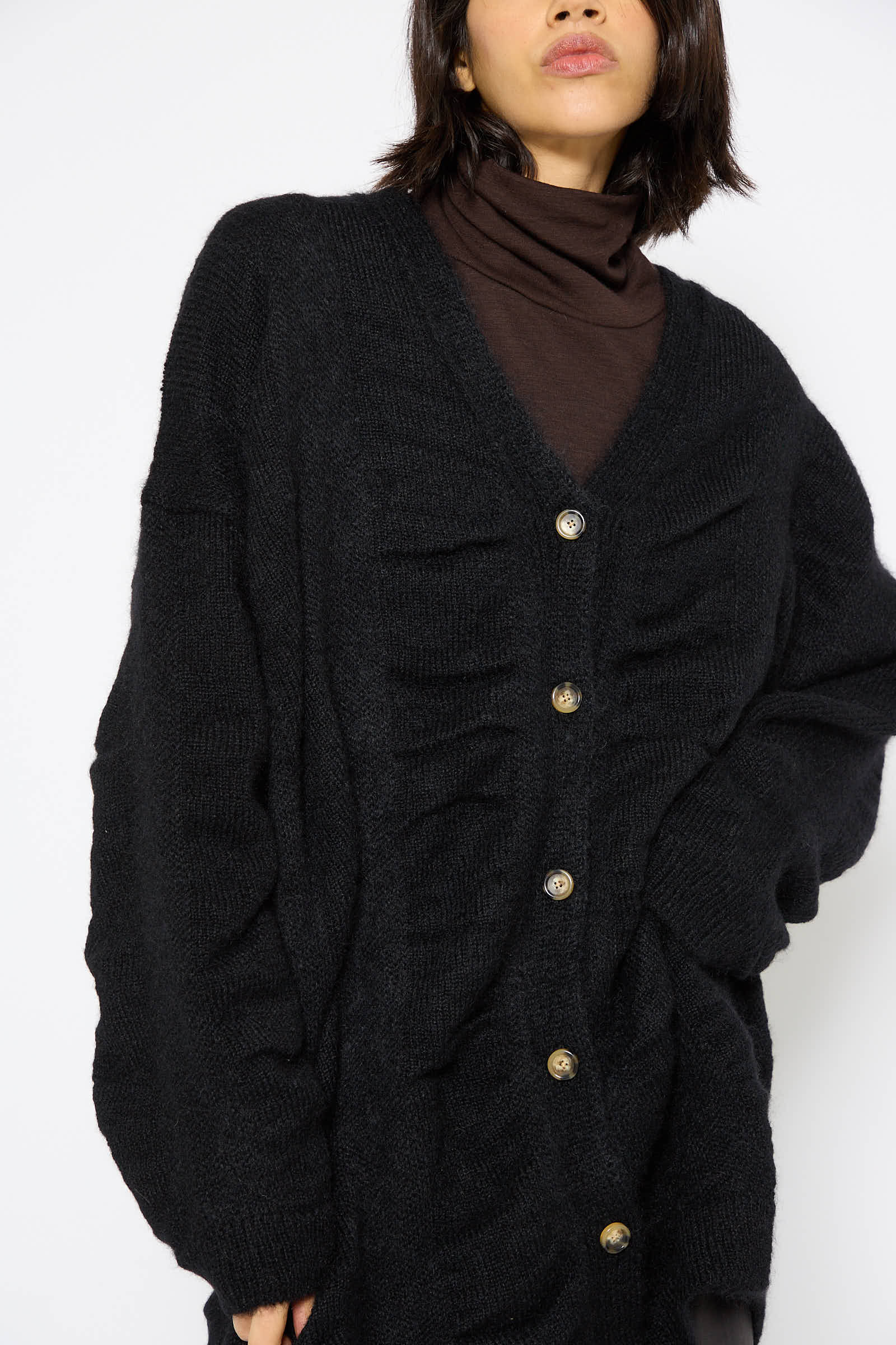 An individual with dark chin-length hair stands gracefully against a plain background, wearing a dark brown turtleneck sweater layered under Black Crane's Japanese Mohair and Wool Waterfall Cardigan in Black. The cardigan, featuring an oversized fit and buttoned-up front, exudes cozy elegance.