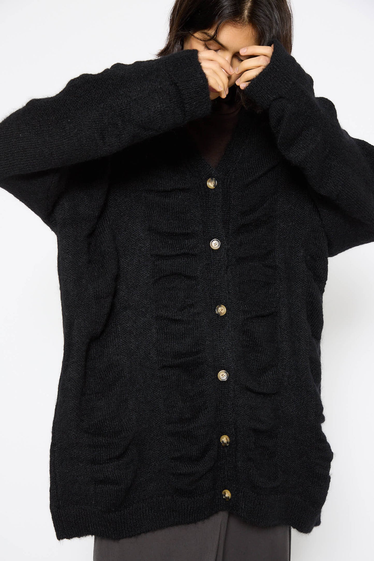 A person wearing the Black Crane Japanese Mohair and Wool Waterfall Cardigan in Black, featuring button closures and textured detailing, stands against a white background, partially covering their face with their hands.