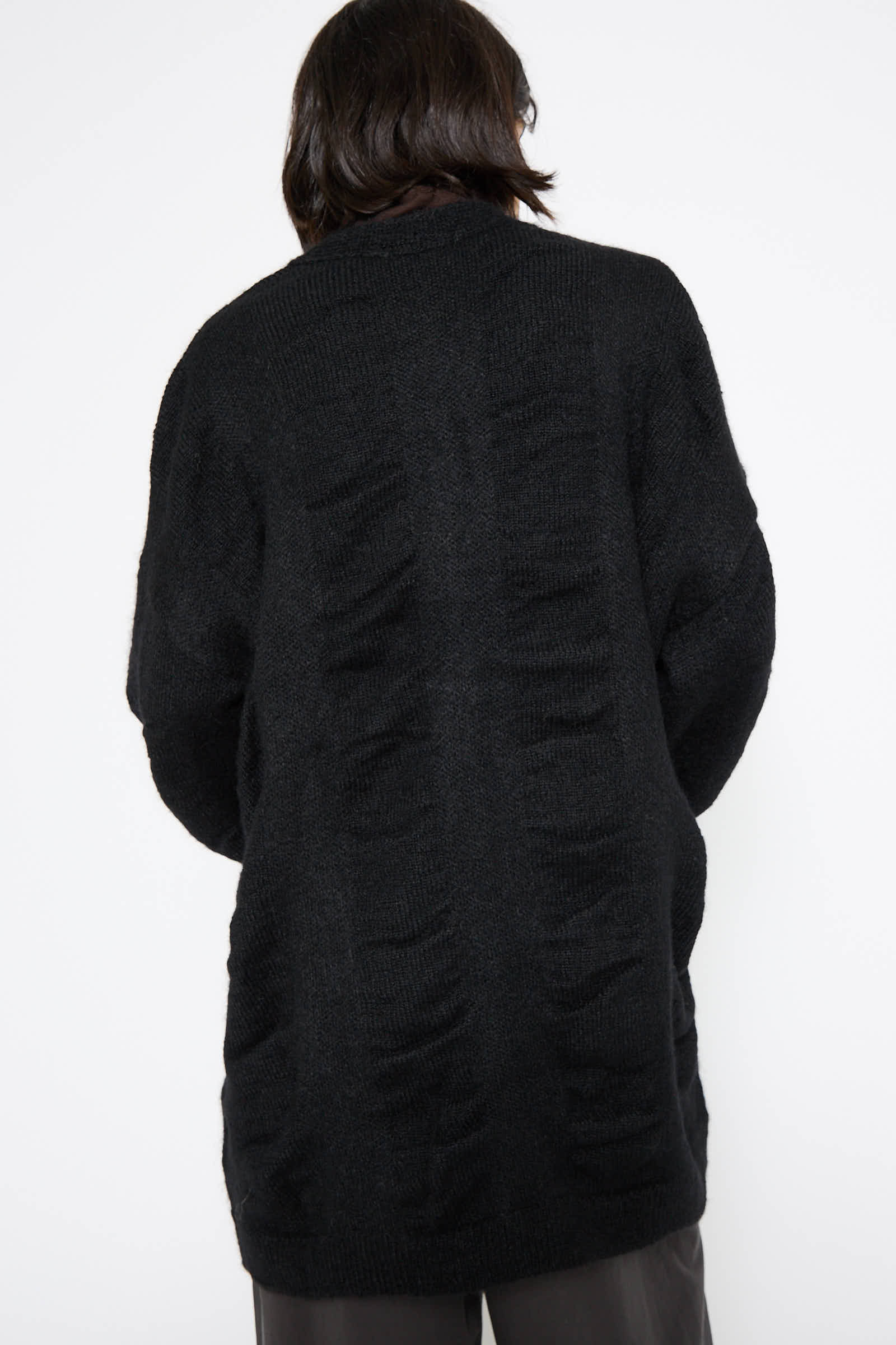 Person in dark clothing with their back turned, wearing the Black Crane Japanese Mohair and Wool Waterfall Cardigan in black and dark pants against a plain background.