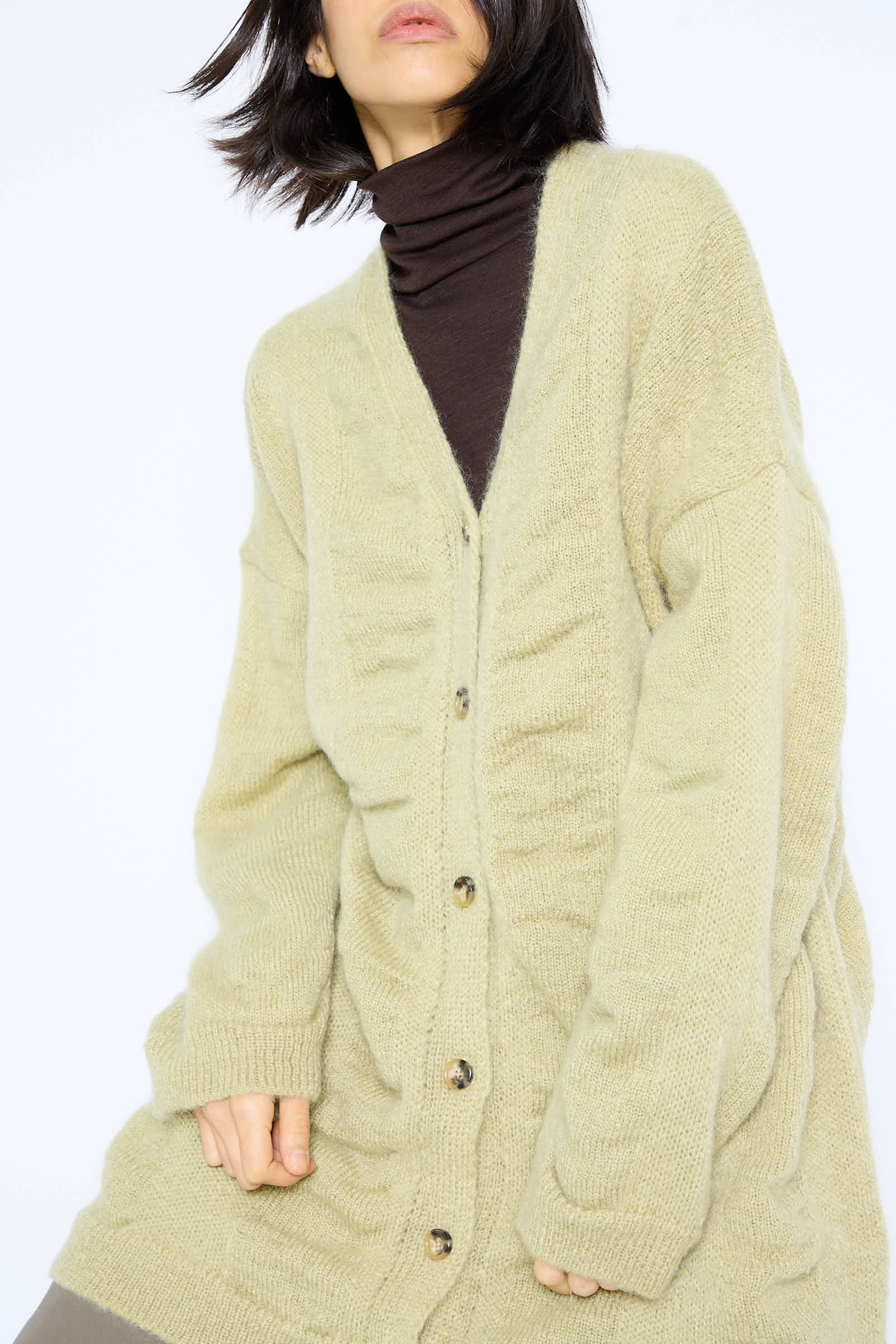 The individual is adorned in a lime-hued, oversized Japanese Mohair and Wool Waterfall Long Cardigan by Black Crane, paired with a brown turtleneck, and is turned slightly to the side.