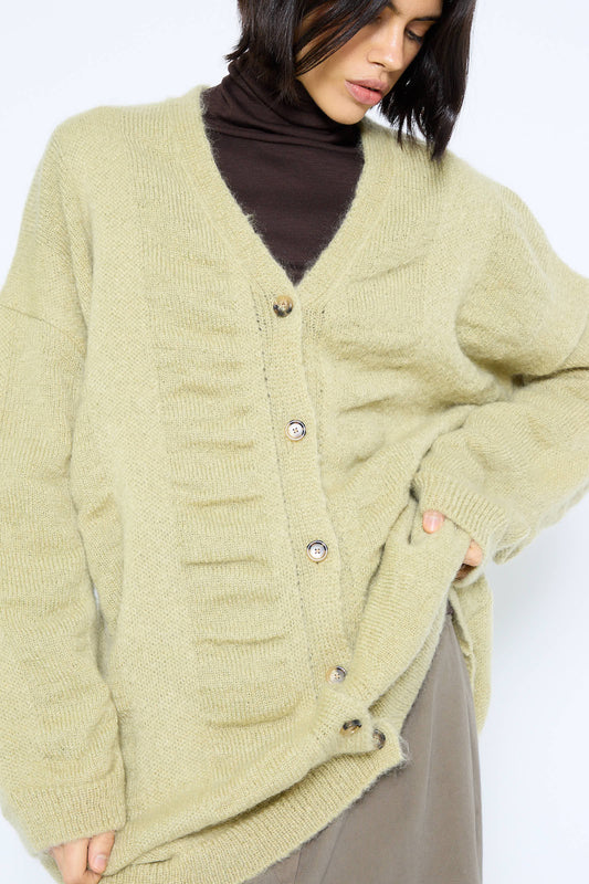 A person wearing a Black Crane Japanese Mohair and Wool Waterfall Long Cardigan in Lime with a button closure over a dark turtleneck top and grey skirt.