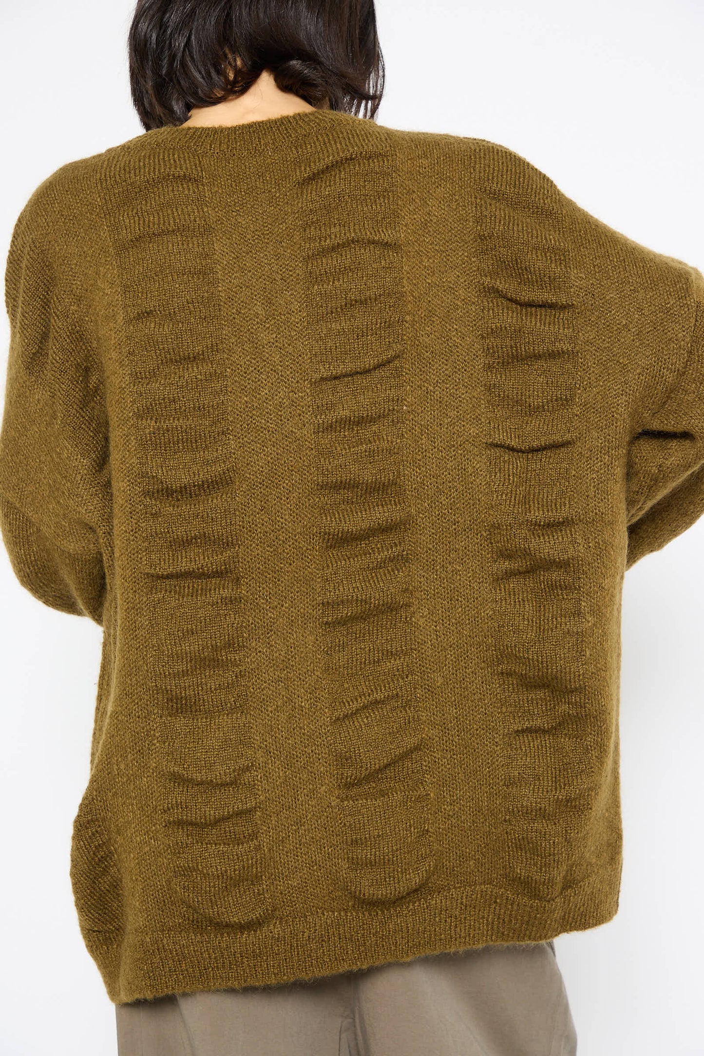 A person is shown from the back wearing the Black Crane Japanese Mohair and Wool Waterfall Sweater in Dark Olive. The oversized sweater, made from a soft Japanese mohair blend, features a textured pattern, has a loose fit, and falls gracefully to the hips.