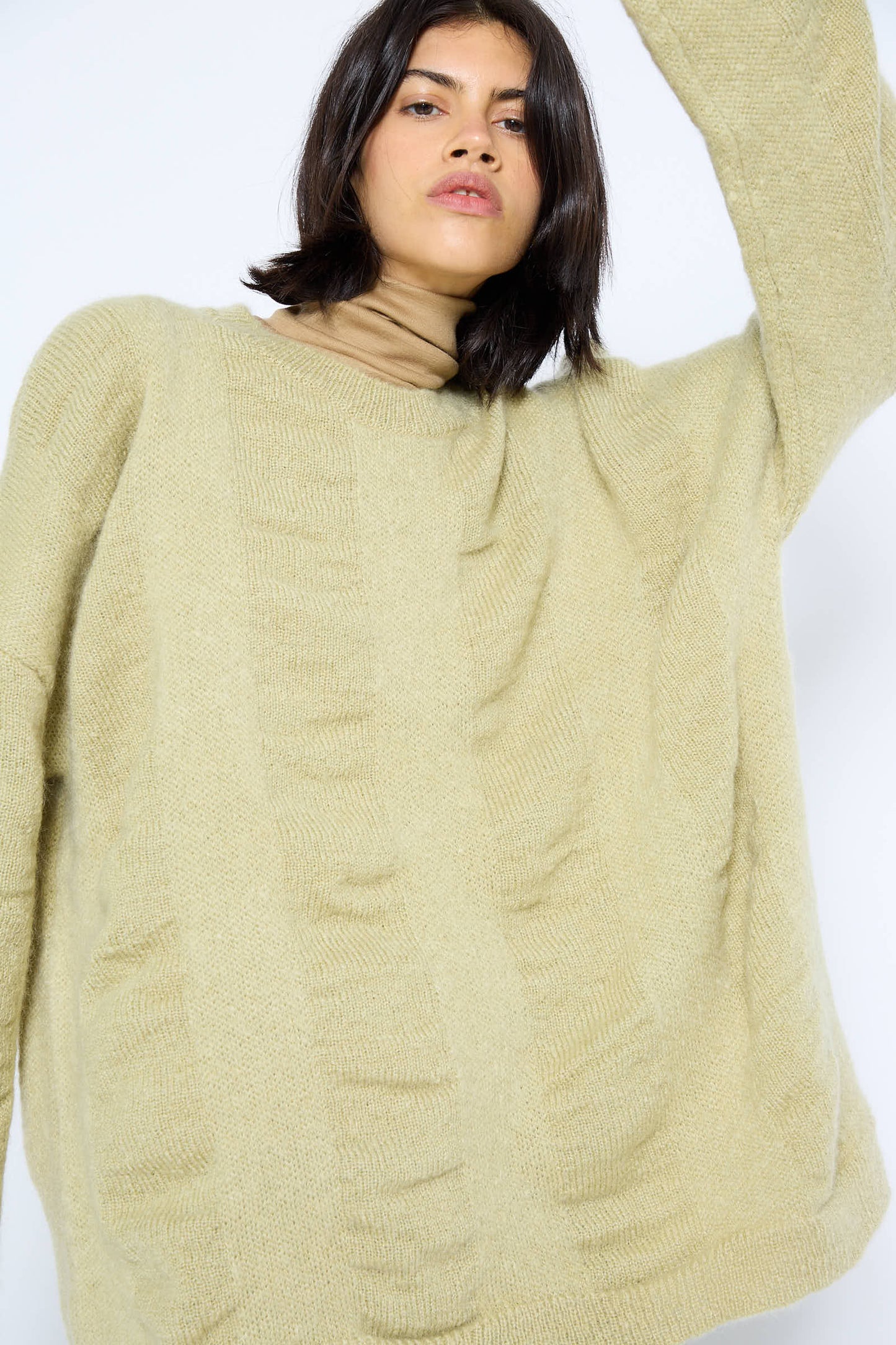 Person wearing the Black Crane Japanese Mohair and Wool Waterfall Sweater in Lime with a high-neck undershirt, raising their right arm. The cozy ensemble features a luxurious mohair blend, adding softness to the look.