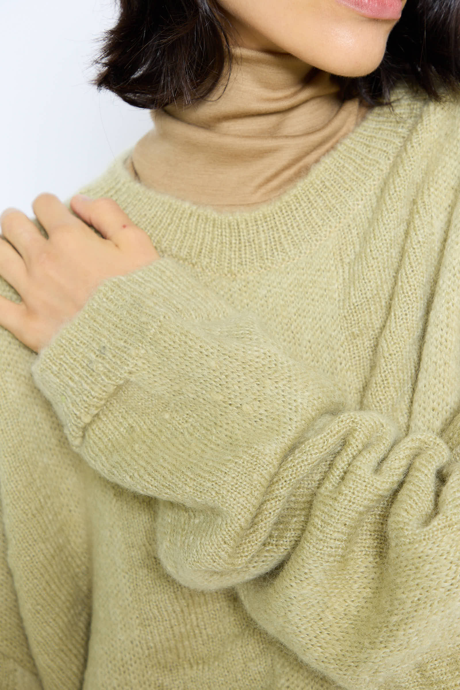 A person wearing an oversized, beige sweater layered over the Black Crane Japanese Mohair and Wool Waterfall Sweater in Lime, with one hand resting on their shoulder.