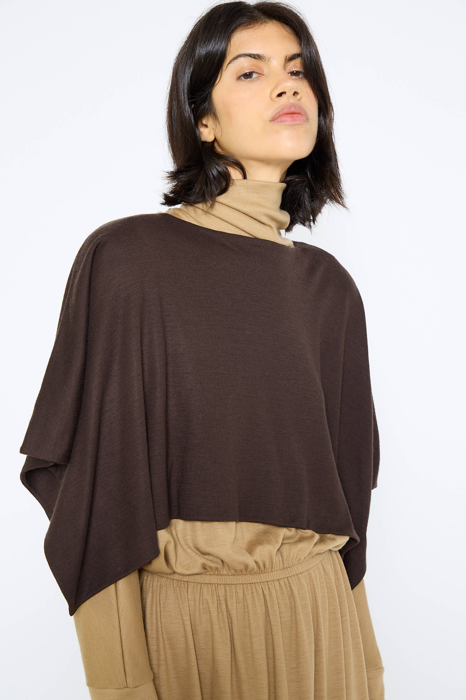 A person wearing a drapey fit, Japanese Wool Jersey Mini Poncho in Cacao by Black Crane over a beige turtleneck and beige high-waisted pants, standing and looking straight ahead.