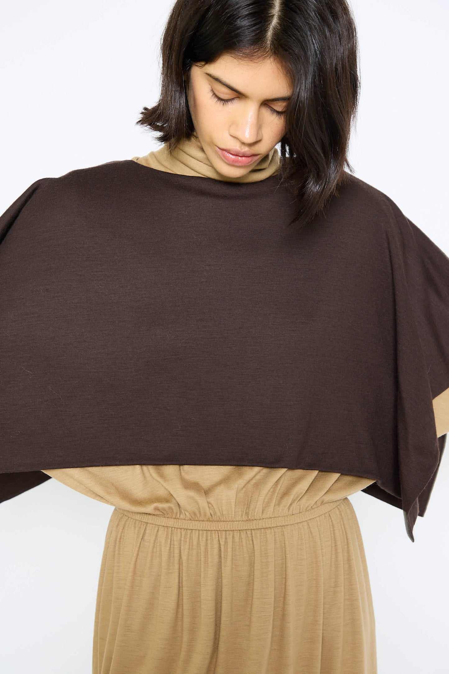 A person wearing a Black Crane Japanese Wool Jersey Mini Poncho in Cacao looks downward.