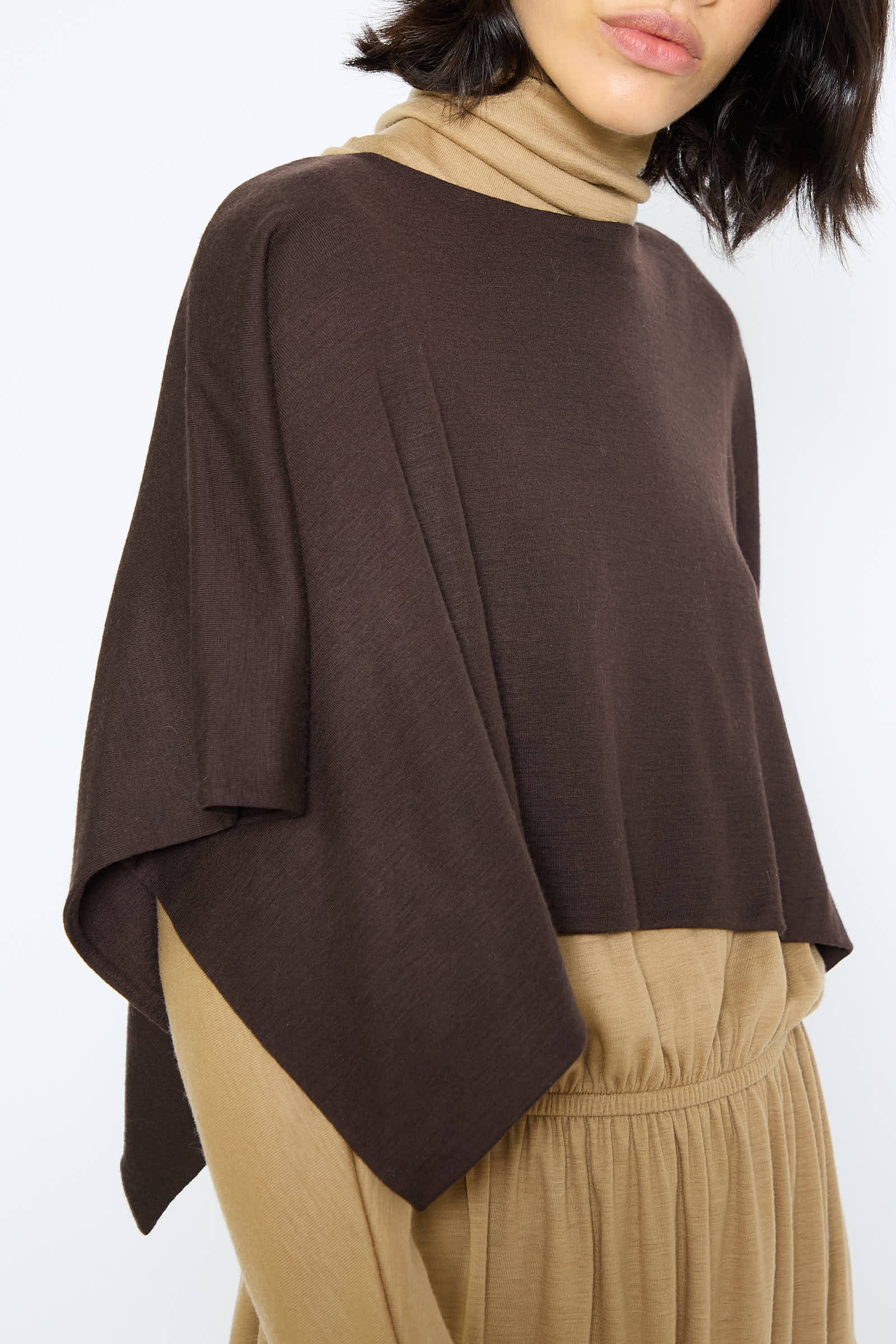 A person in a Japanese Wool Jersey Mini Poncho in Cacao by Black Crane, stylishly layered over a tan turtleneck dress.