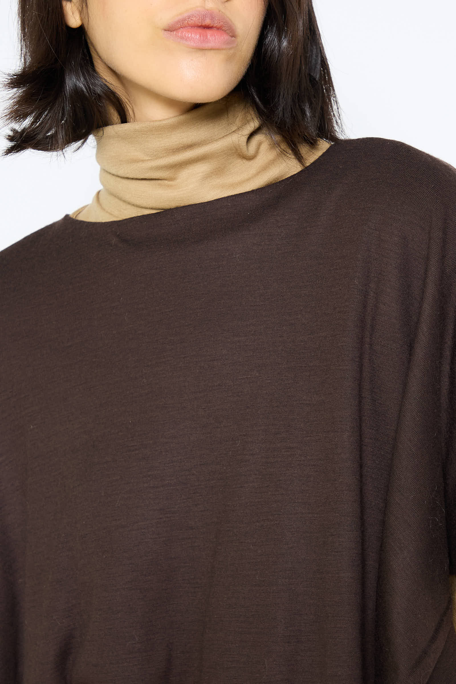 A person with short dark hair, wearing a brown turtleneck over a beige top, is showcased from the shoulders up, resembling the drapey fit of the Japanese Wool Jersey Mini Poncho in Cacao by Black Crane.