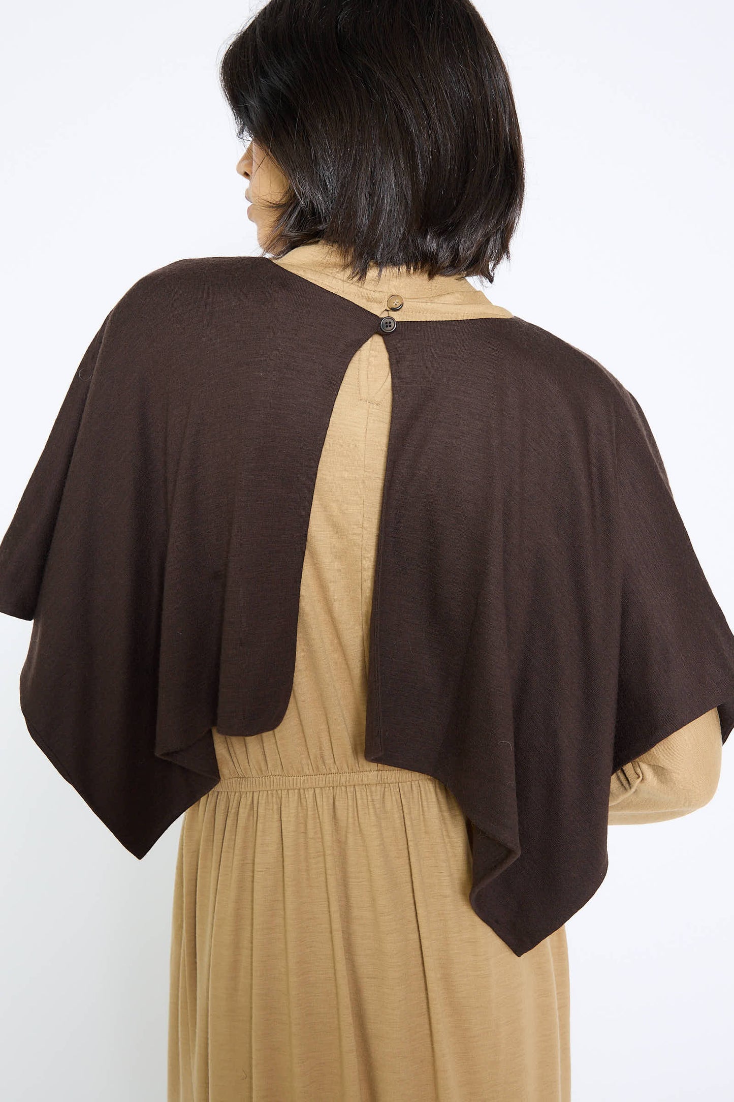 A person with short, dark hair is shown from behind. They are wearing a tan dress complemented by the Black Crane Japanese Wool Jersey Mini Poncho in Cacao, which features a drapey fit and is fastened at the back with two buttons.