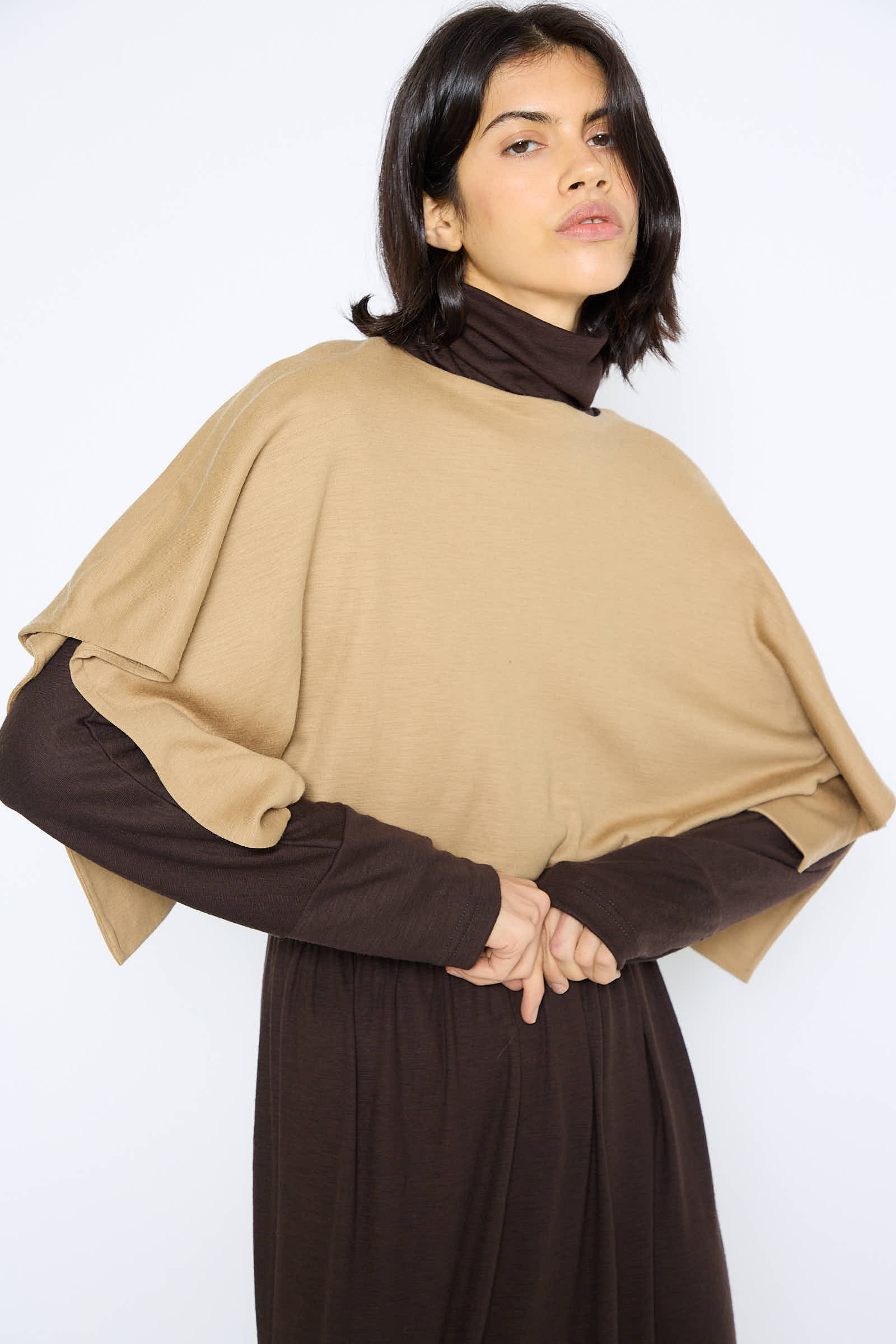 A person with shoulder-length dark hair wearing the Black Crane Japanese Wool Jersey Mini Poncho in Camel over a brown turtleneck stands against a white background.