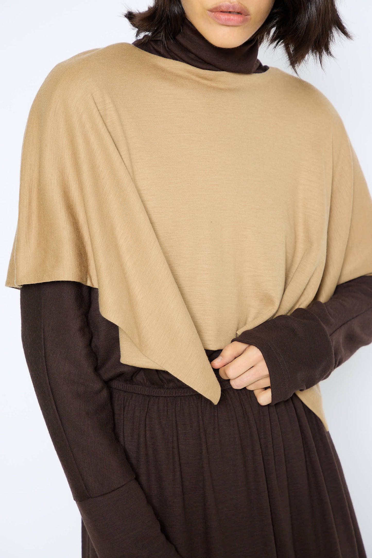 A person wearing the Black Crane Japanese Wool Jersey Mini Poncho in Camel, paired with a dark brown long-sleeve garment underneath, with one hand resting on their waist.