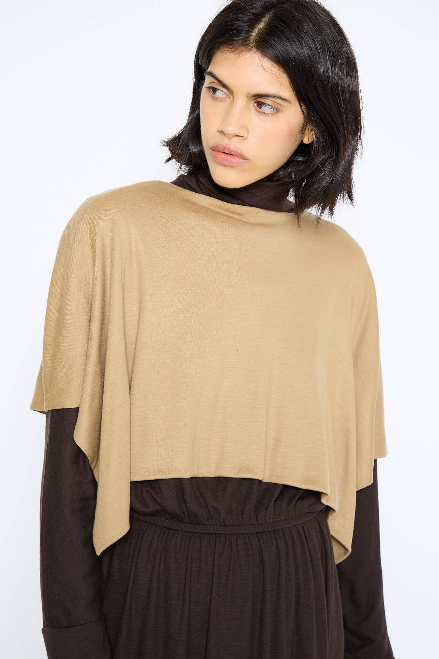 A person with medium-length dark hair wears the Black Crane Japanese Wool Jersey Mini Poncho in Camel over a dark brown long-sleeve top and matching high-waisted pants, against a white background.