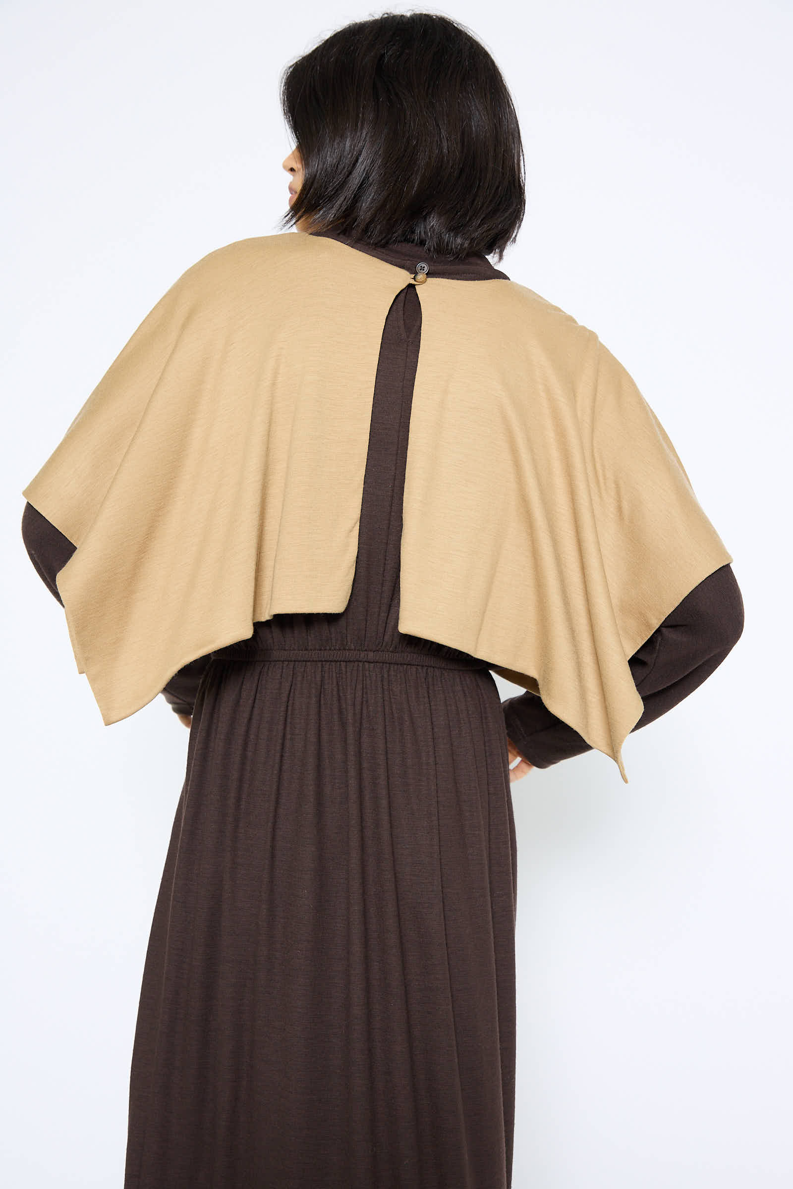 A person wearing a long, dark brown dress paired with the Japanese Wool Jersey Mini Poncho in Camel by Black Crane stands with their back to the camera, showcasing the drapey fit and intricate back design of the outfit.