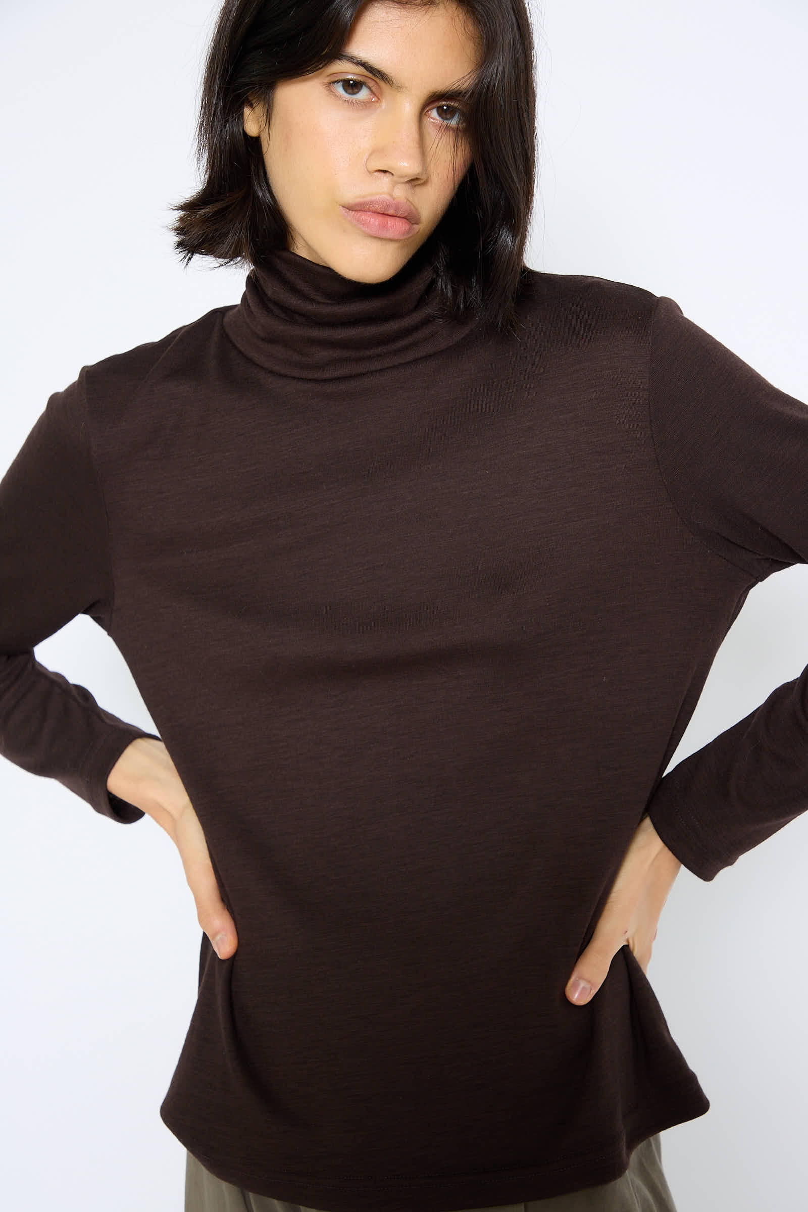 A person with short dark hair wearing a Black Crane Japanese Wool Jersey Turtleneck in Cacao stands with hands on hips against a plain background.