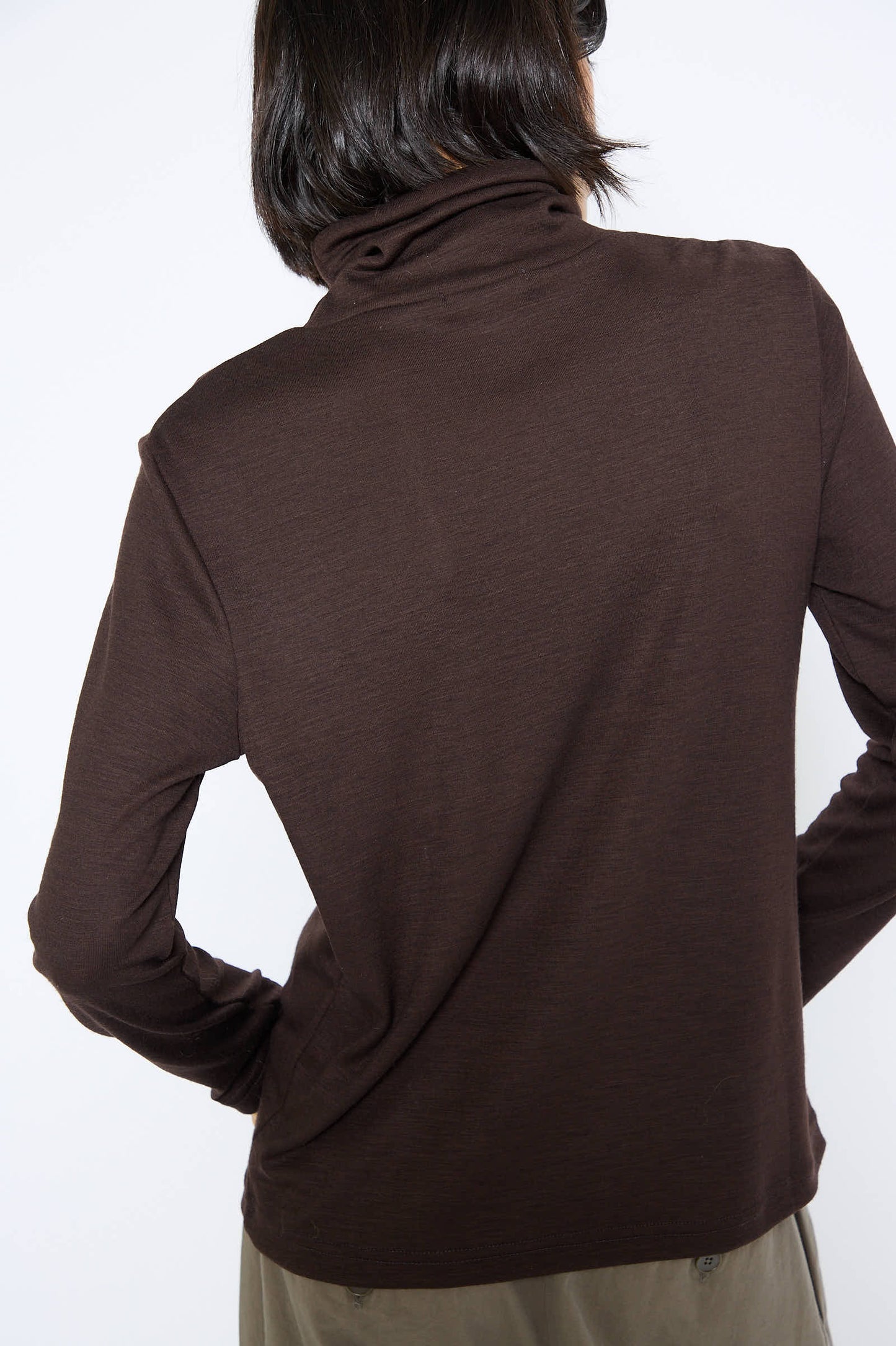 A person with shoulder-length dark hair is shown from the back, wearing a long-sleeved Japanese Wool Jersey Turtleneck in Cacao by Black Crane.