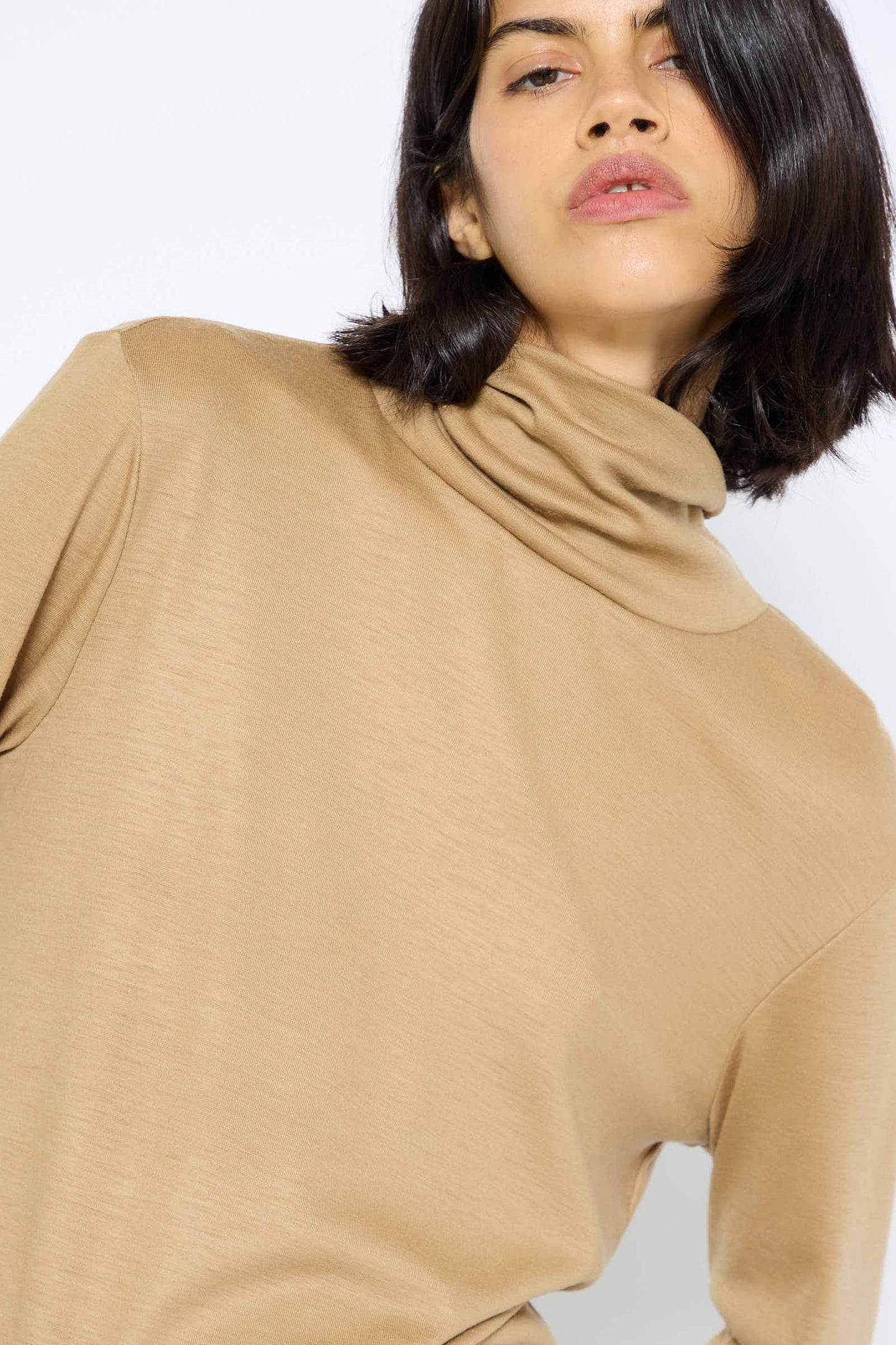 A person with shoulder-length dark hair, wearing a Japanese Wool Jersey Turtleneck in Camel by Black Crane, looks slightly upwards.