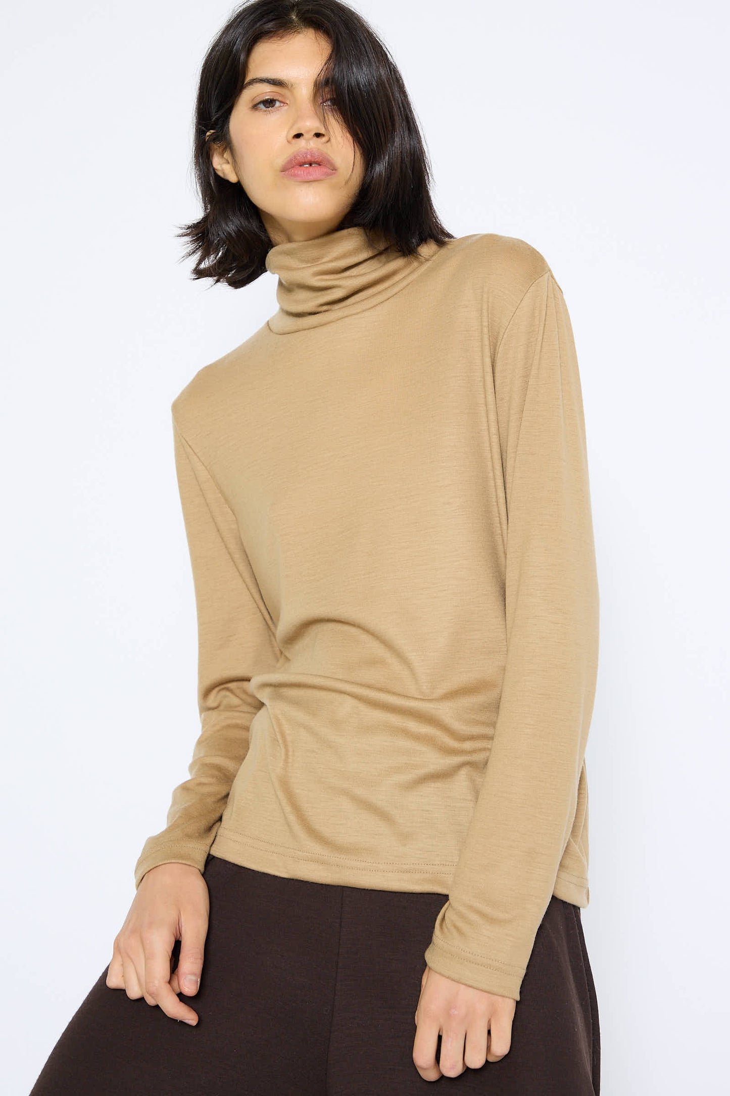 A person with shoulder-length dark hair wearing the Black Crane Japanese Wool Jersey Turtleneck in Camel and dark pants poses against a white background.