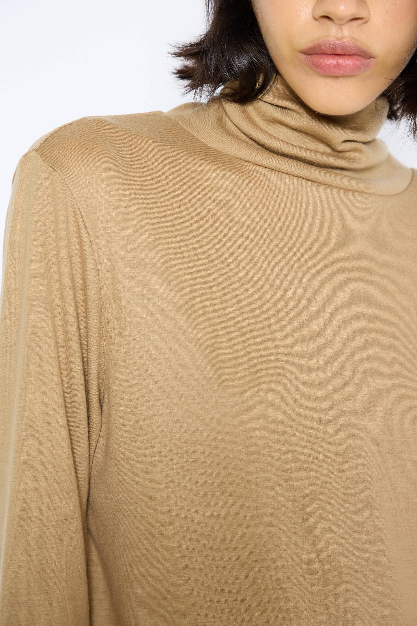 A close-up image showcases a person wearing the Black Crane Japanese Wool Jersey Turtleneck in Camel. The image captures only the lower half of their face and neck area, highlighting the fine texture of the wool jersey. The person sports short, dark hair.