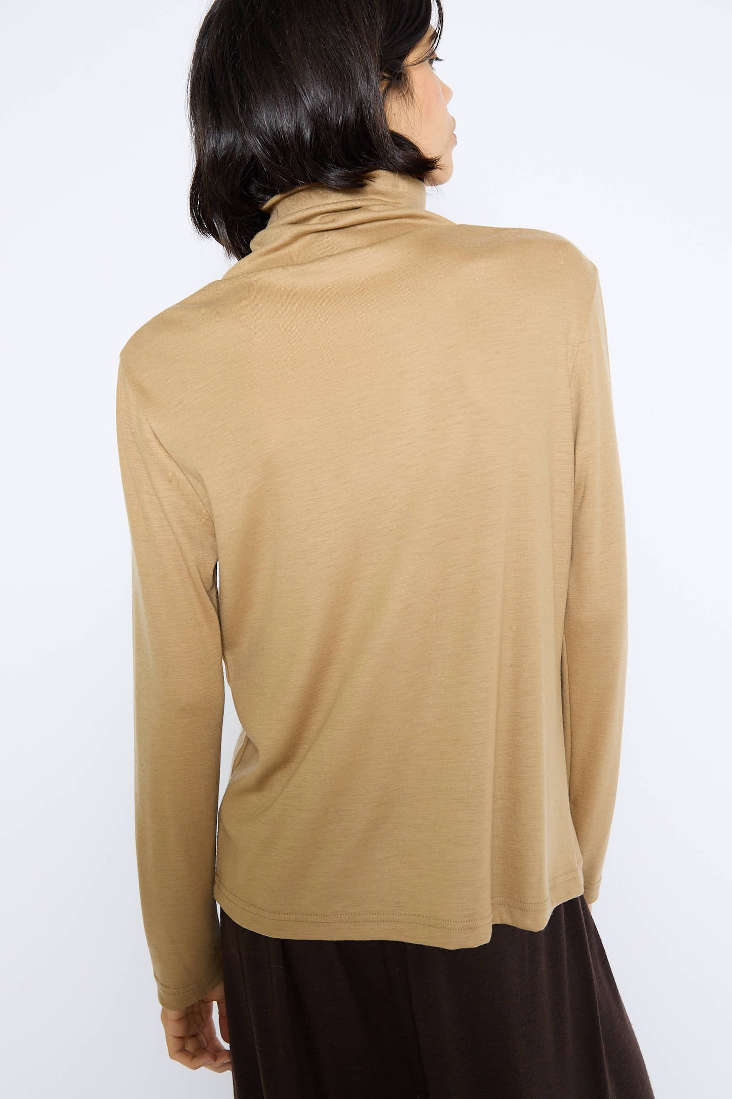 A person with shoulder-length, dark hair is standing with their back to the camera, wearing a relaxed fit, Japanese Wool Jersey Turtleneck in Camel by Black Crane and dark pants.