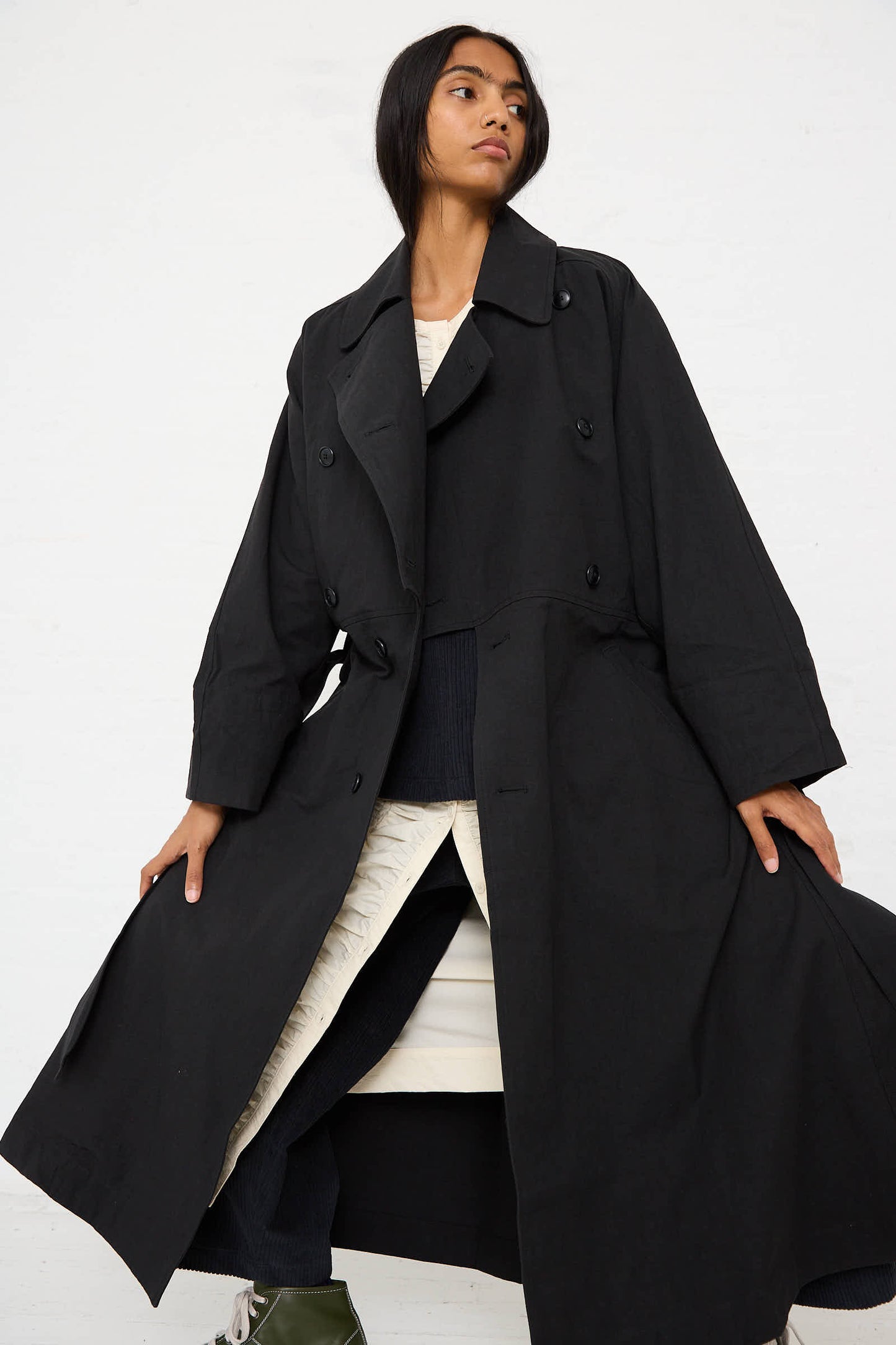 Person in a Woven Cotton Long Coat in Black by Black Crane, layered over a relaxed fit beige outfit, standing confidently against a plain white background.