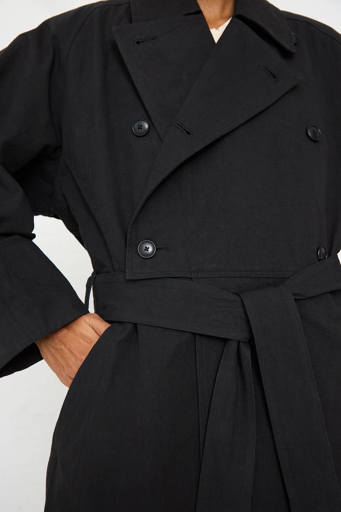 A person wearing the Woven Cotton Long Coat in Black by Black Crane, a black, double-breasted trench coat made from Japanese cotton with a belt tied at the waist, has one hand in their pocket.