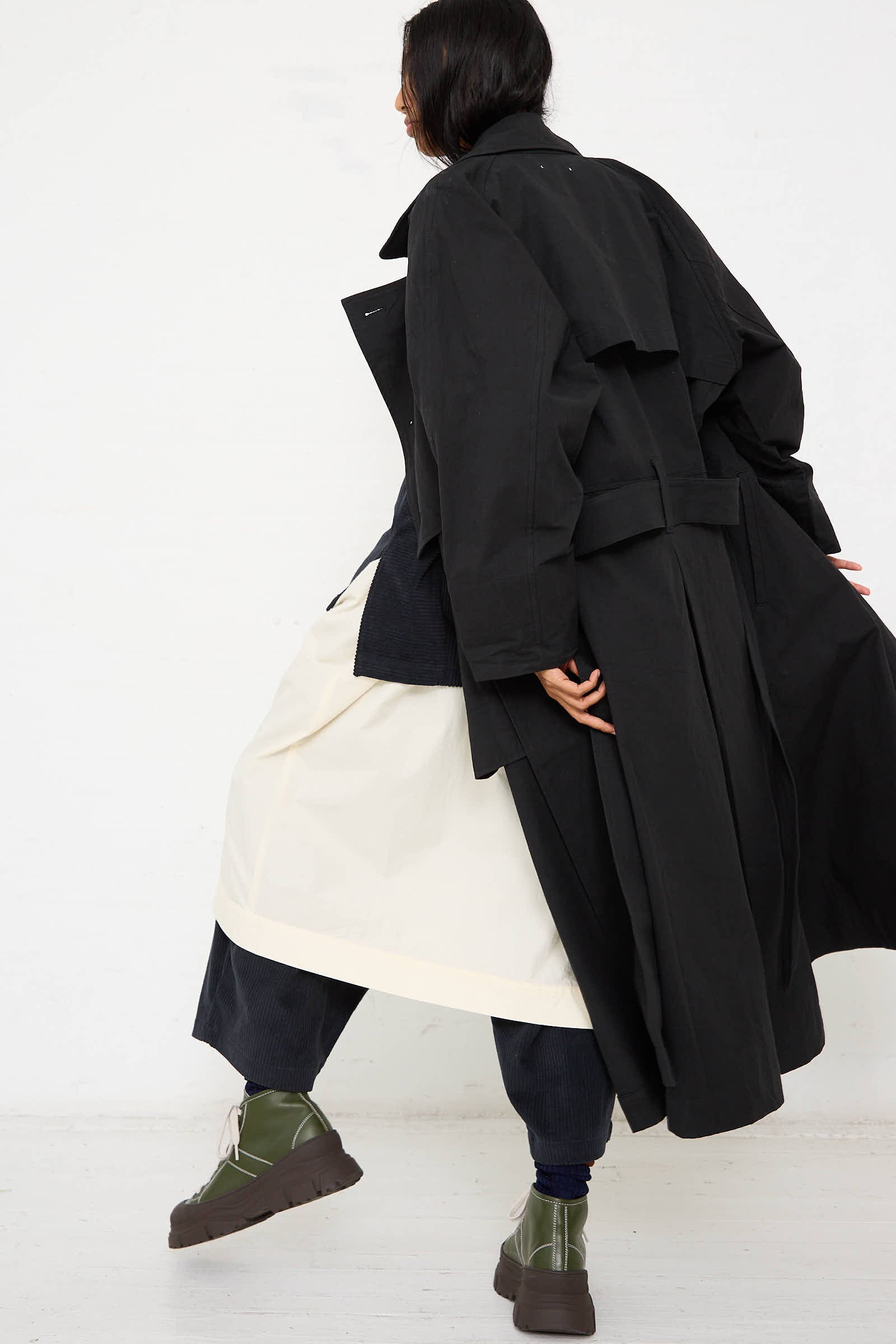 The person wearing the Black Crane Woven Cotton Long Coat in black, paired with a cream-colored skirt and green boots, is turned away while holding the coat. The relaxed fit of the coat showcases the high quality of the Japanese cotton material. The background is plain white.