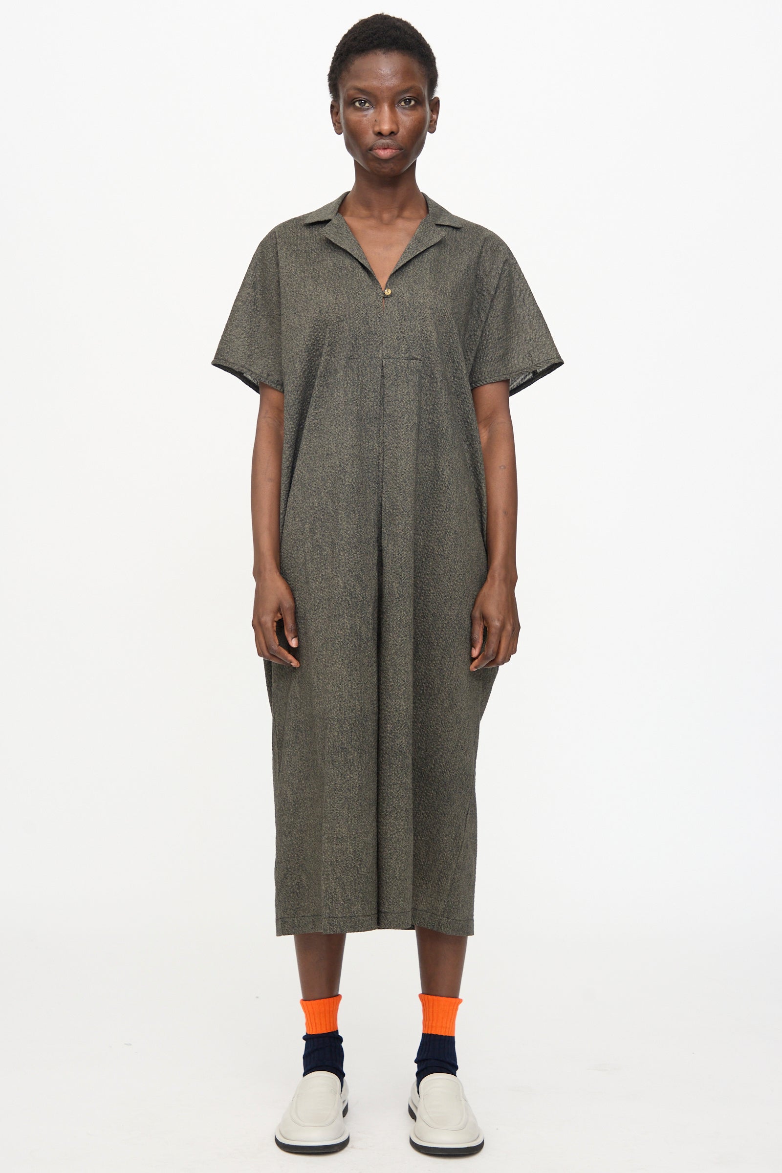 A person stands against a white background wearing the Black Crane Kite Dress with Micro Wave Texture in Steel, styled with orange socks, navy socks, and white slip-on shoes.