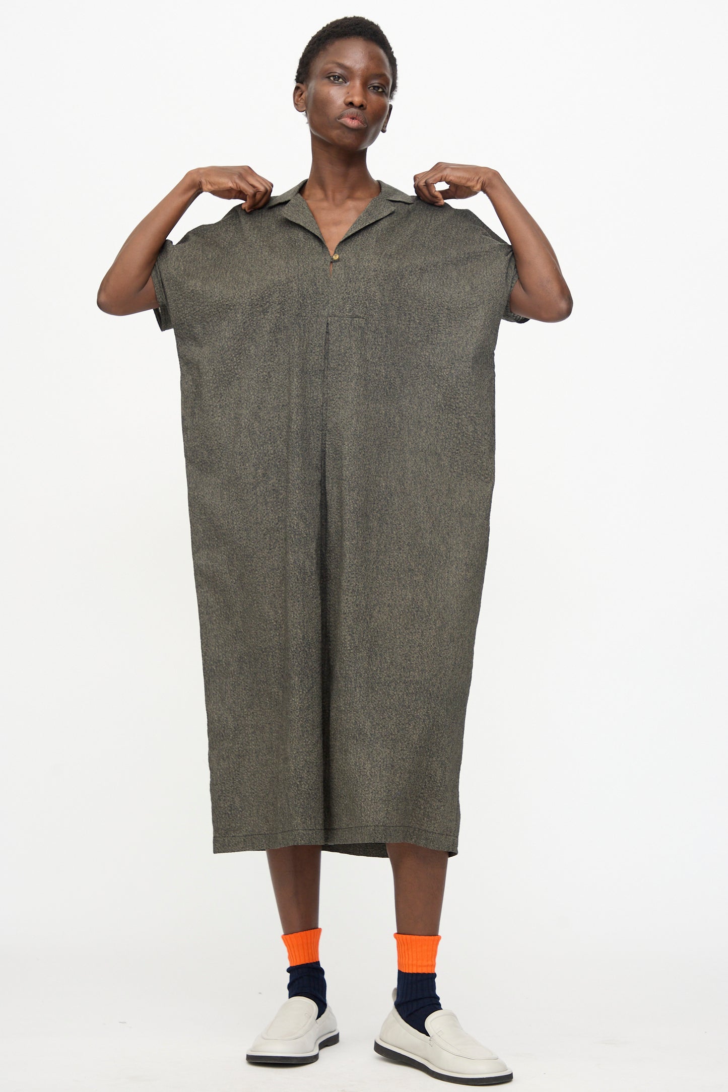 A person stands against a white background wearing the Black Crane Kite Dress with Micro Wave Texture in Steel, paired with orange and dark socks and white slip-on shoes, holding the dress at the shoulders.