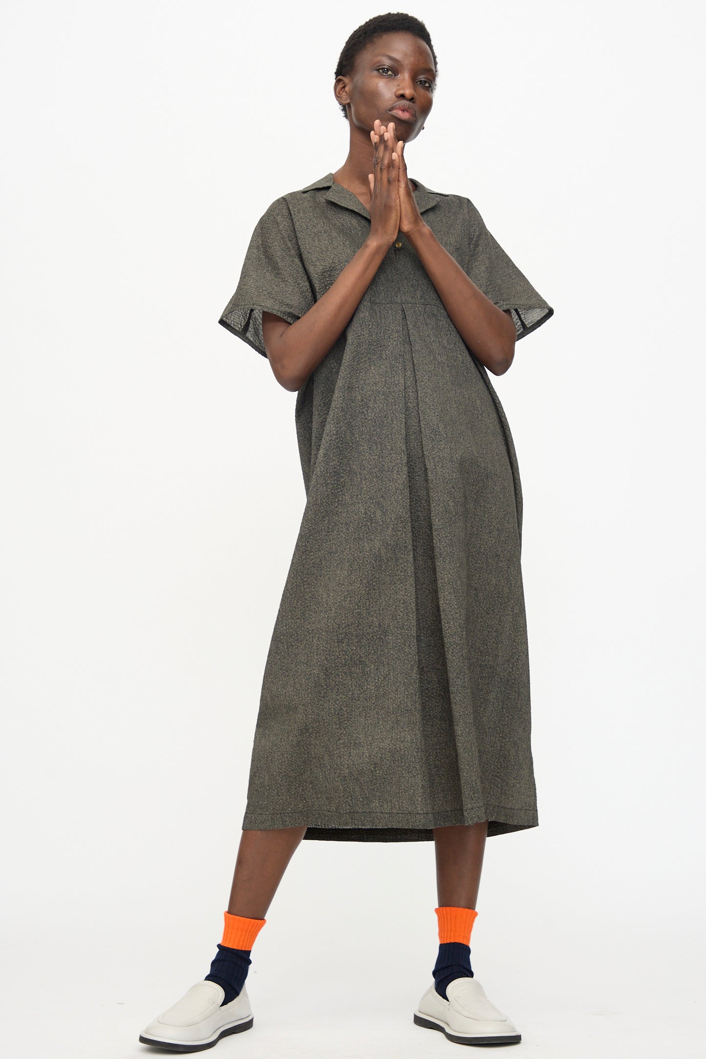 A person stands against a white background wearing the Black Crane Kite Dress with Micro Wave Texture in Steel—relaxed fit and short sleeves—paired with orange socks and white slip-on shoes, hands pressed together near their face.