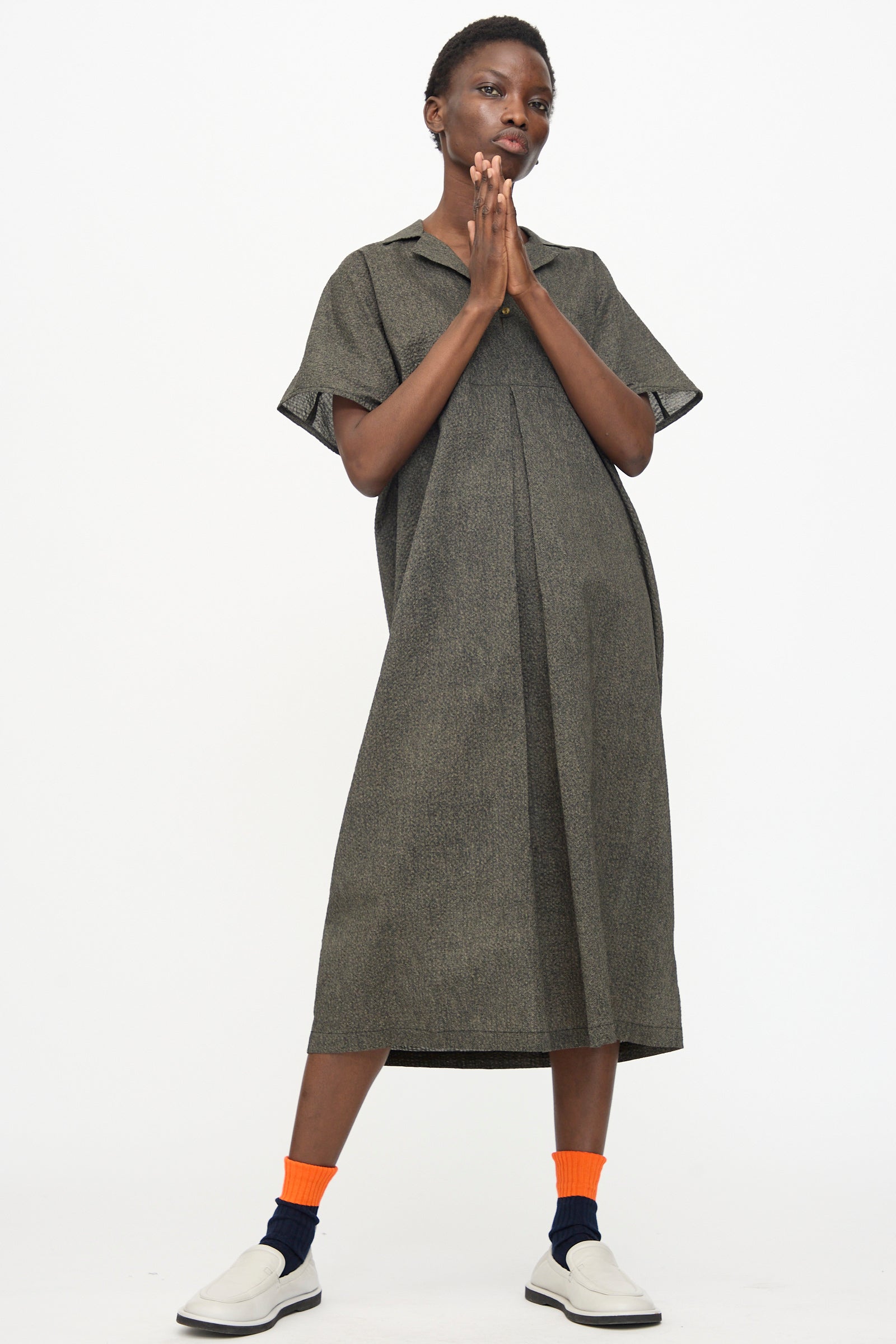 A person stands against a white background wearing the Black Crane Kite Dress with Micro Wave Texture in Steel—relaxed fit and short sleeves—paired with orange socks and white slip-on shoes, hands pressed together near their face.