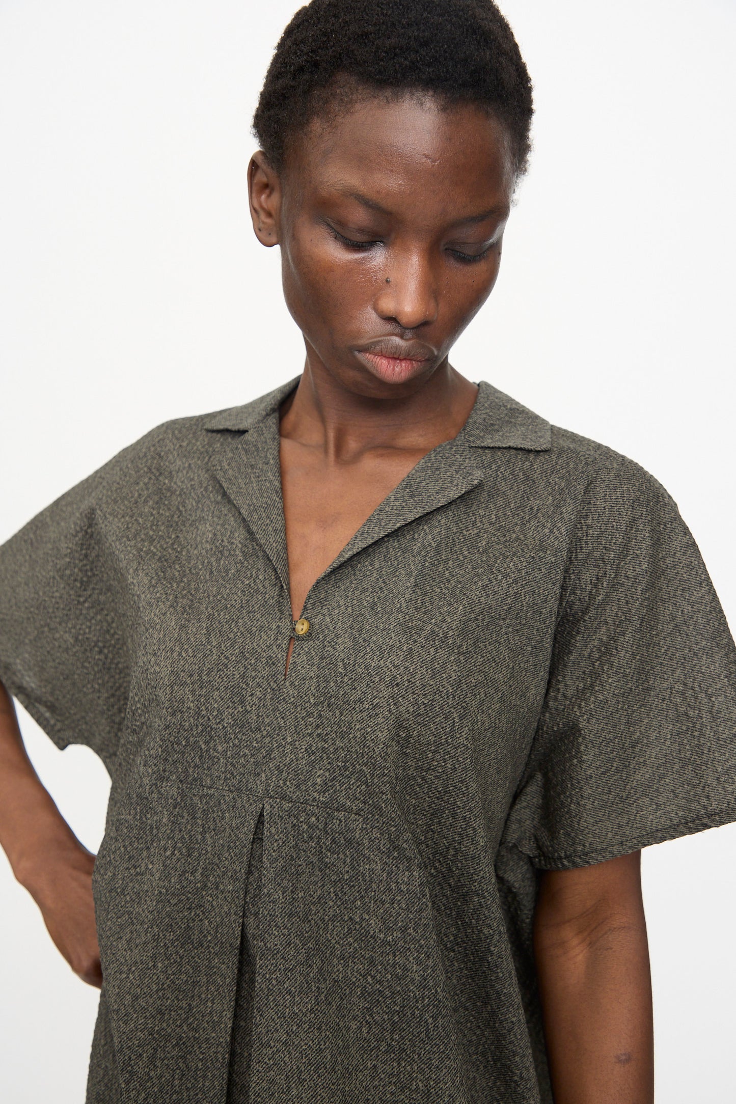 A person wears the Black Crane Kite Dress with Micro Wave Texture in Steel, a relaxed fit, short-sleeve collared dress, standing against a plain white background and looking downward.